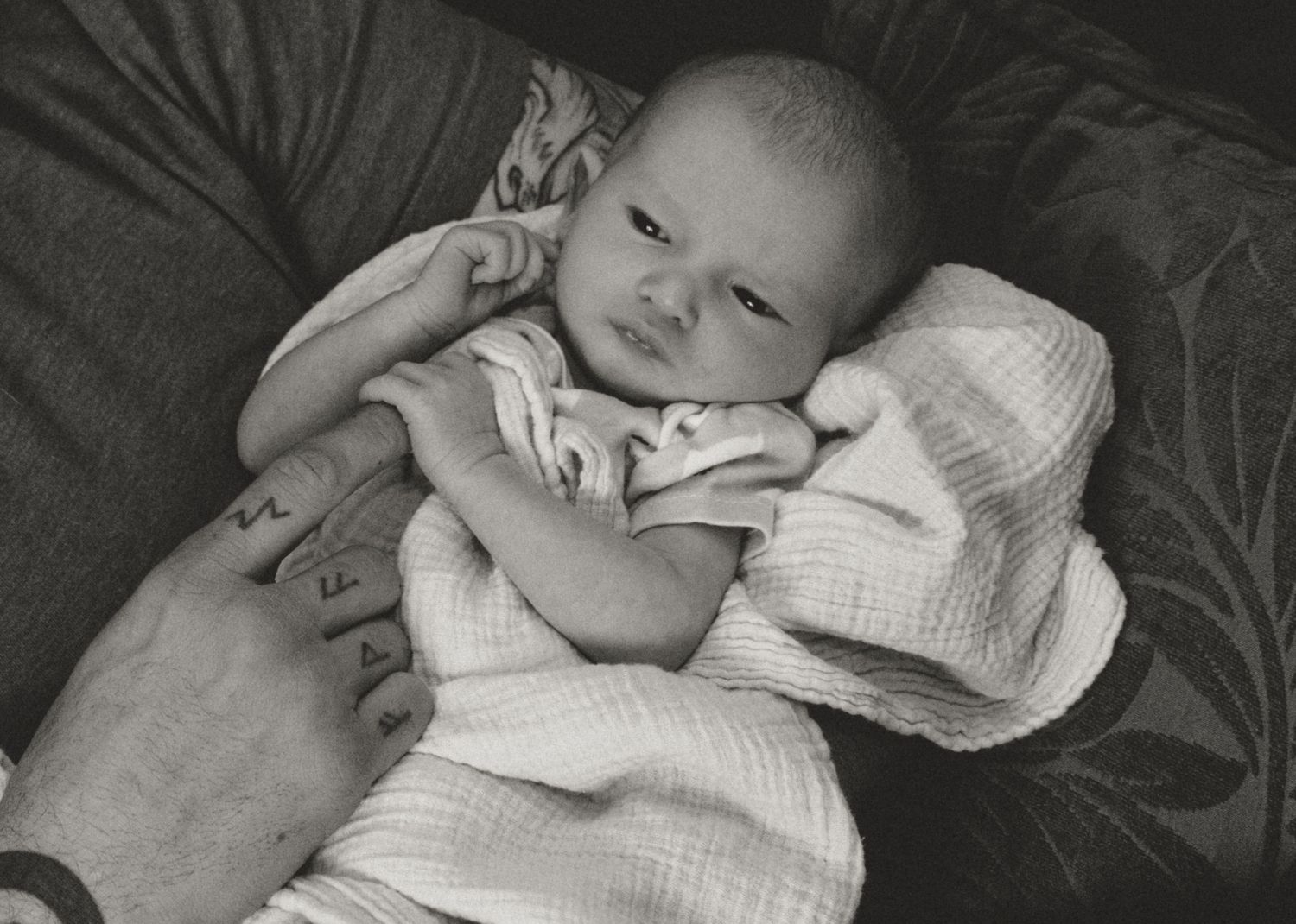 Close-up black and white photo of newborn baby resting on parent's tattooed hand.