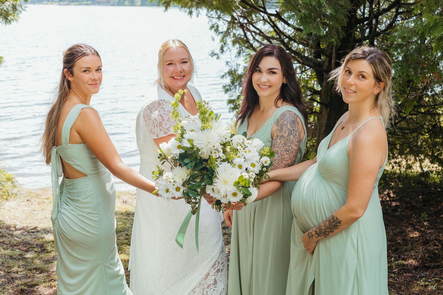 Blonde bride poses with three bridesmaids in sage green dresses holding white bouquets near a lake.