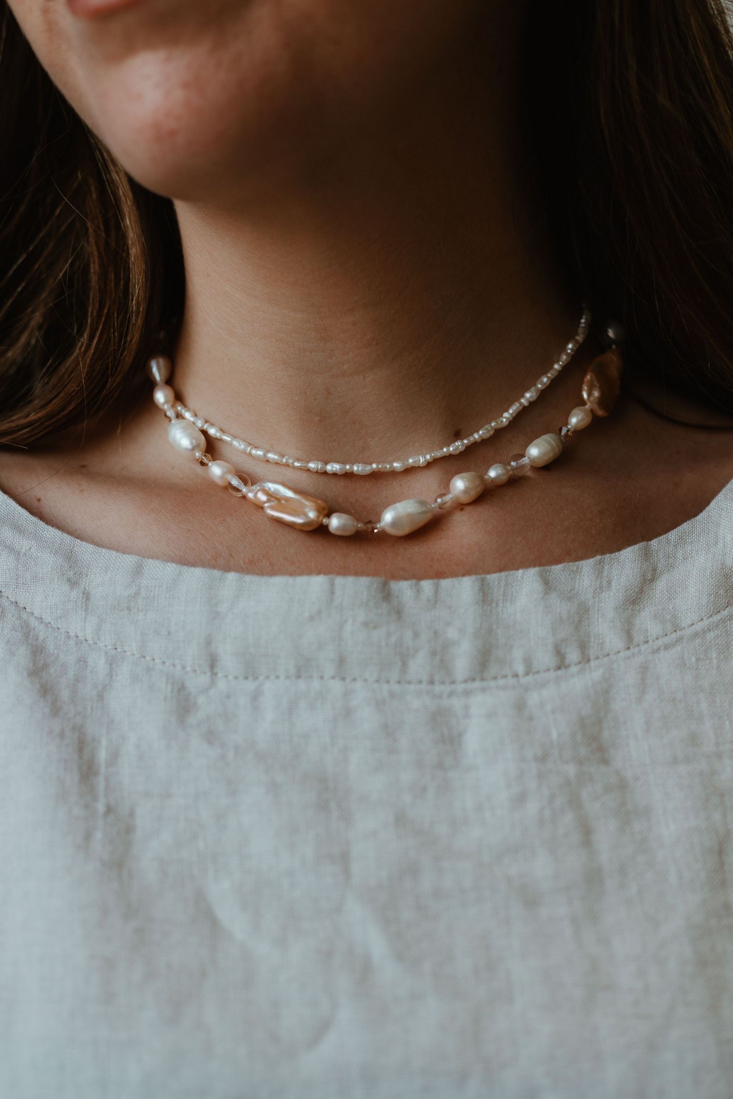 Woman wearing two layered pearl necklaces, featuring baroque and seed pearls, paired with a white linen top.