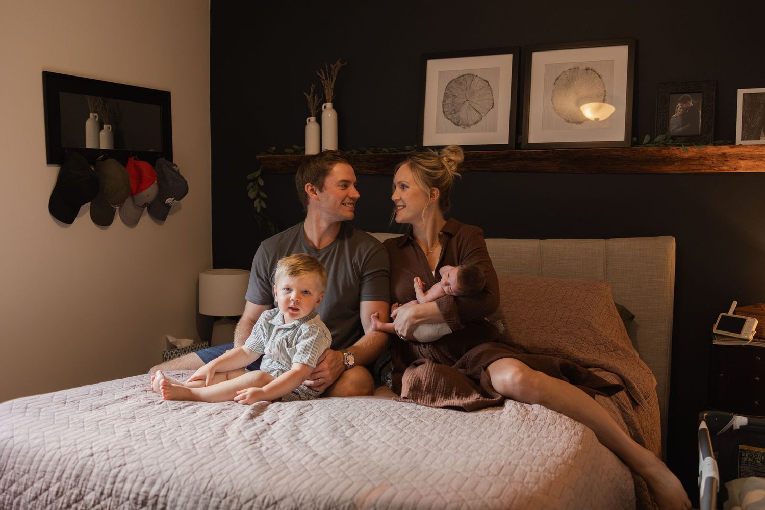 Family of four sitting on a bed, parents smiling with toddler and newborn baby in a cozy bedroom.