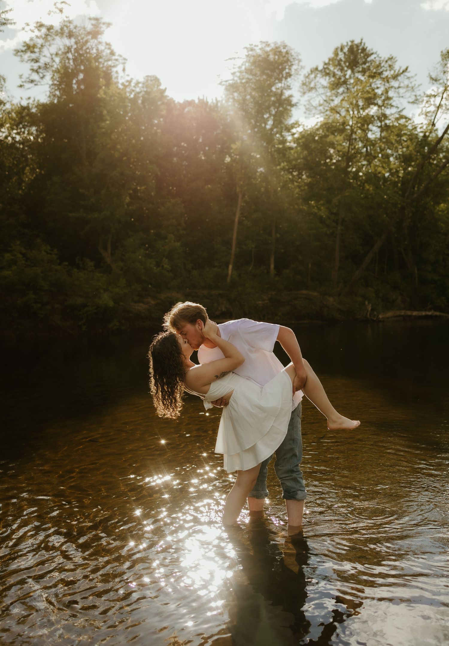 Couple sharing a romantic kiss in a sunlit river, surrounded by lush green trees during golden hour.
