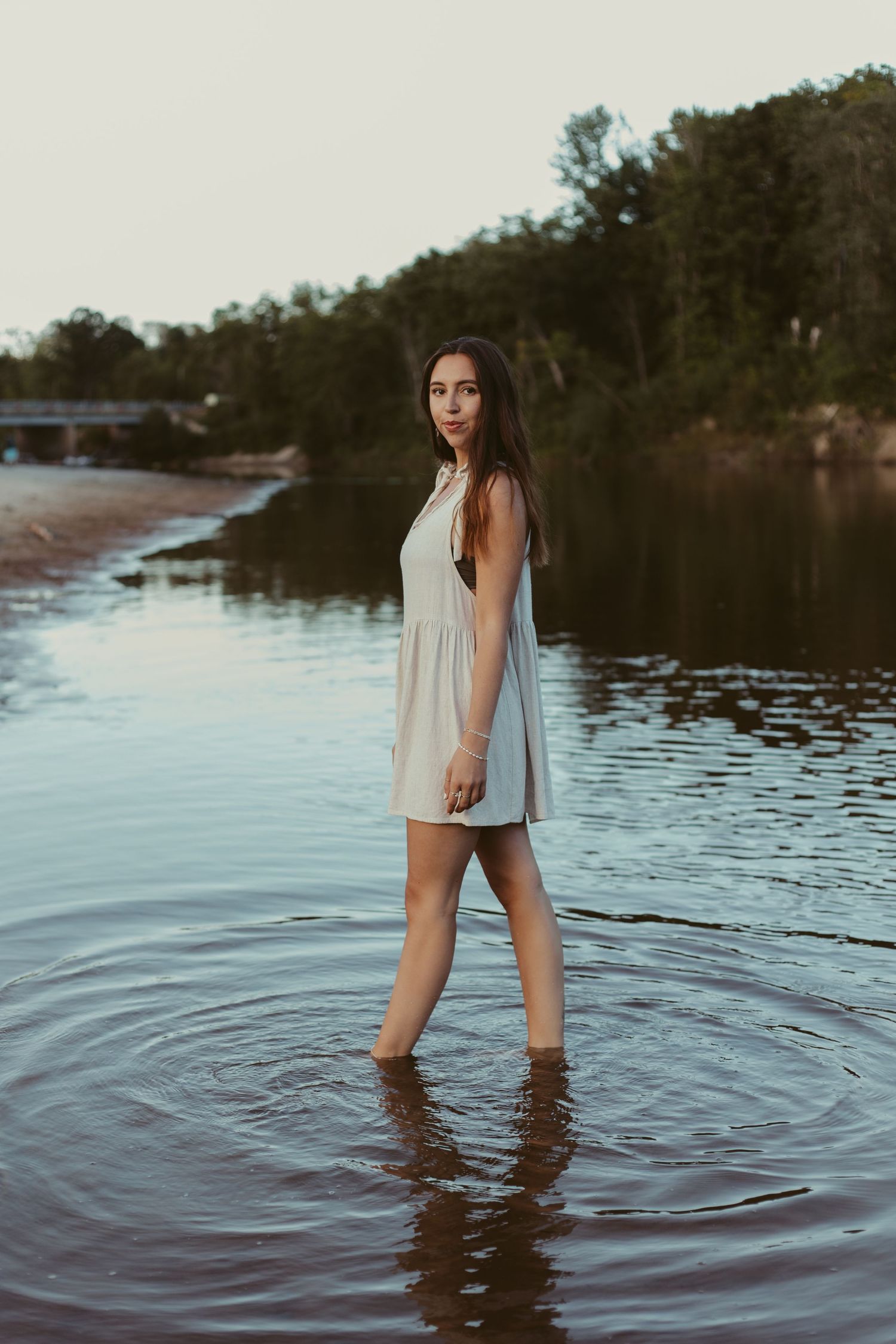 Young woman in white dress wades through calm river at dusk near forest.