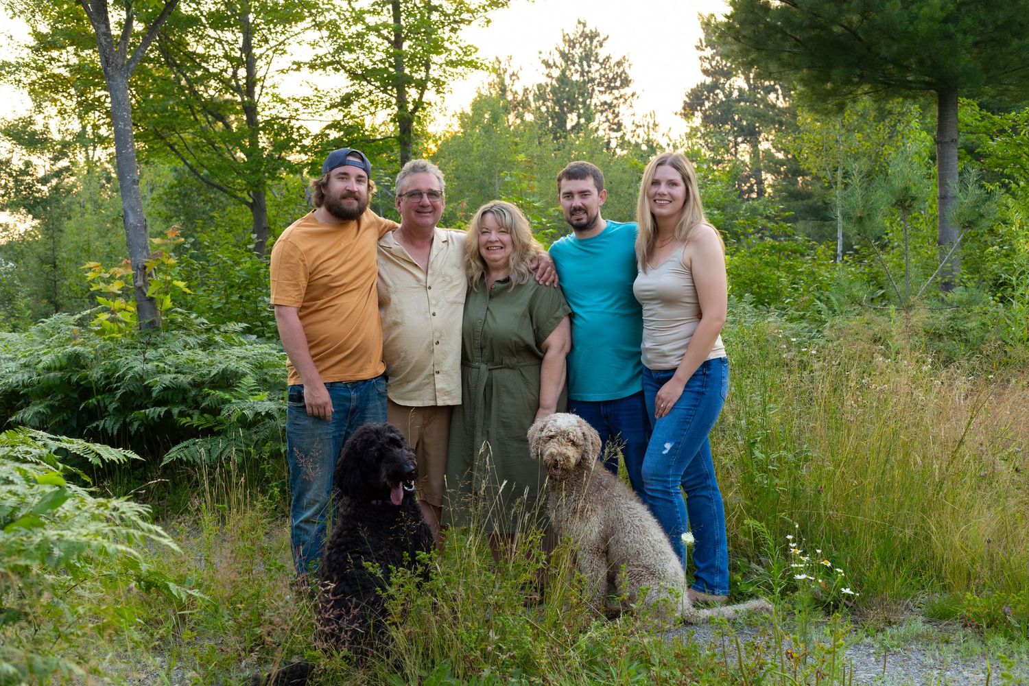 Happy family group photo with two dogs surrounded by tall grass and trees at dusk.