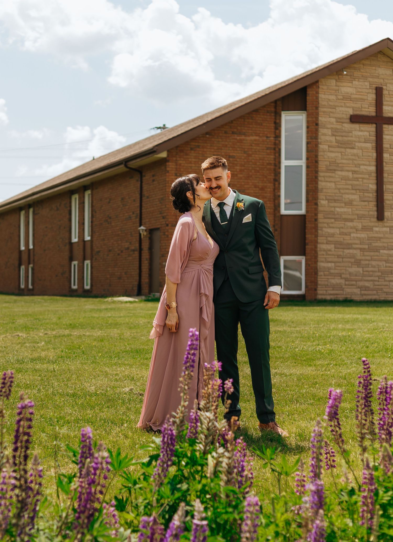 Couple in formal attire standing in front of a brick church with a cross, surrounded by purple lupine flowers.