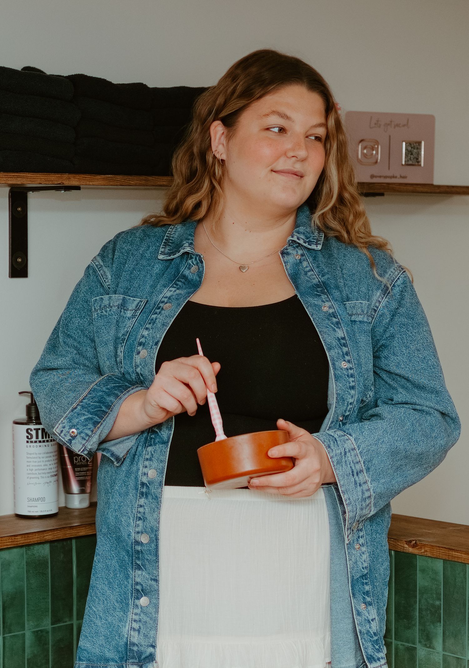 Woman in denim jacket holding a wooden bowl with a pink spoon, standing near green tiled wall.