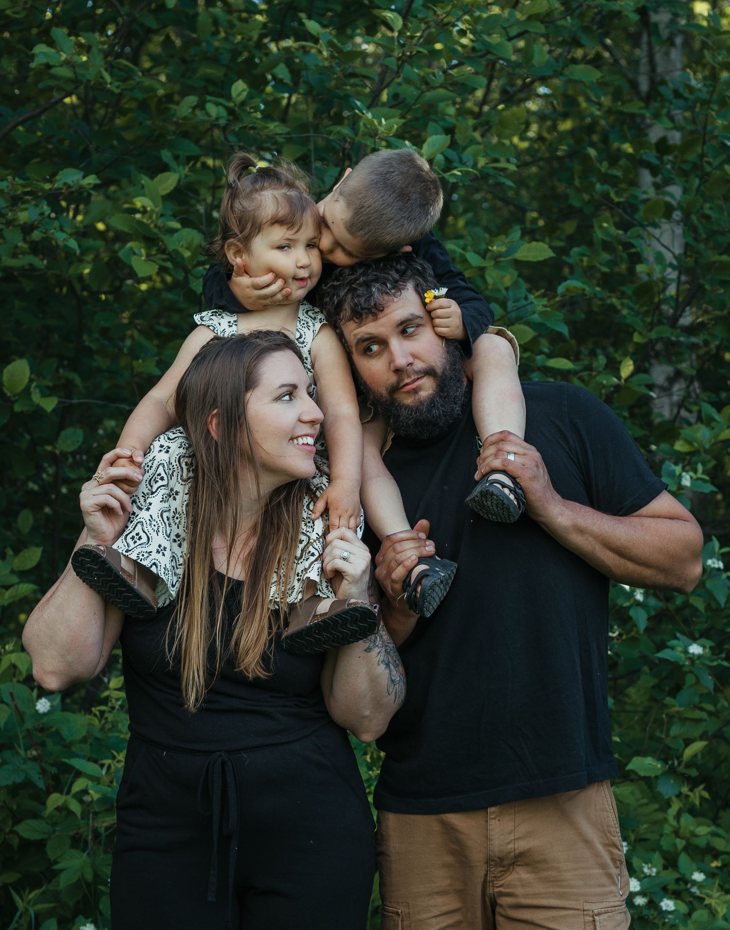 A young family of four poses outdoors, with two parents each holding a child among lush green trees.