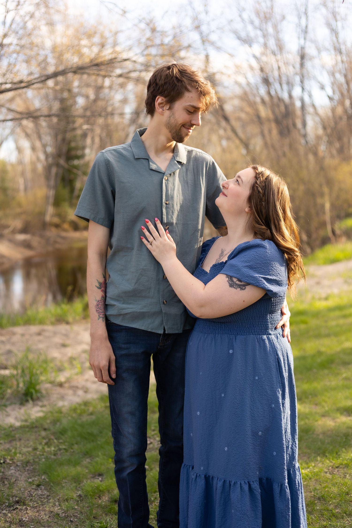 A couple smiles at each other outdoors, woman in blue dress with hand on mans chest, trees and pond in background.