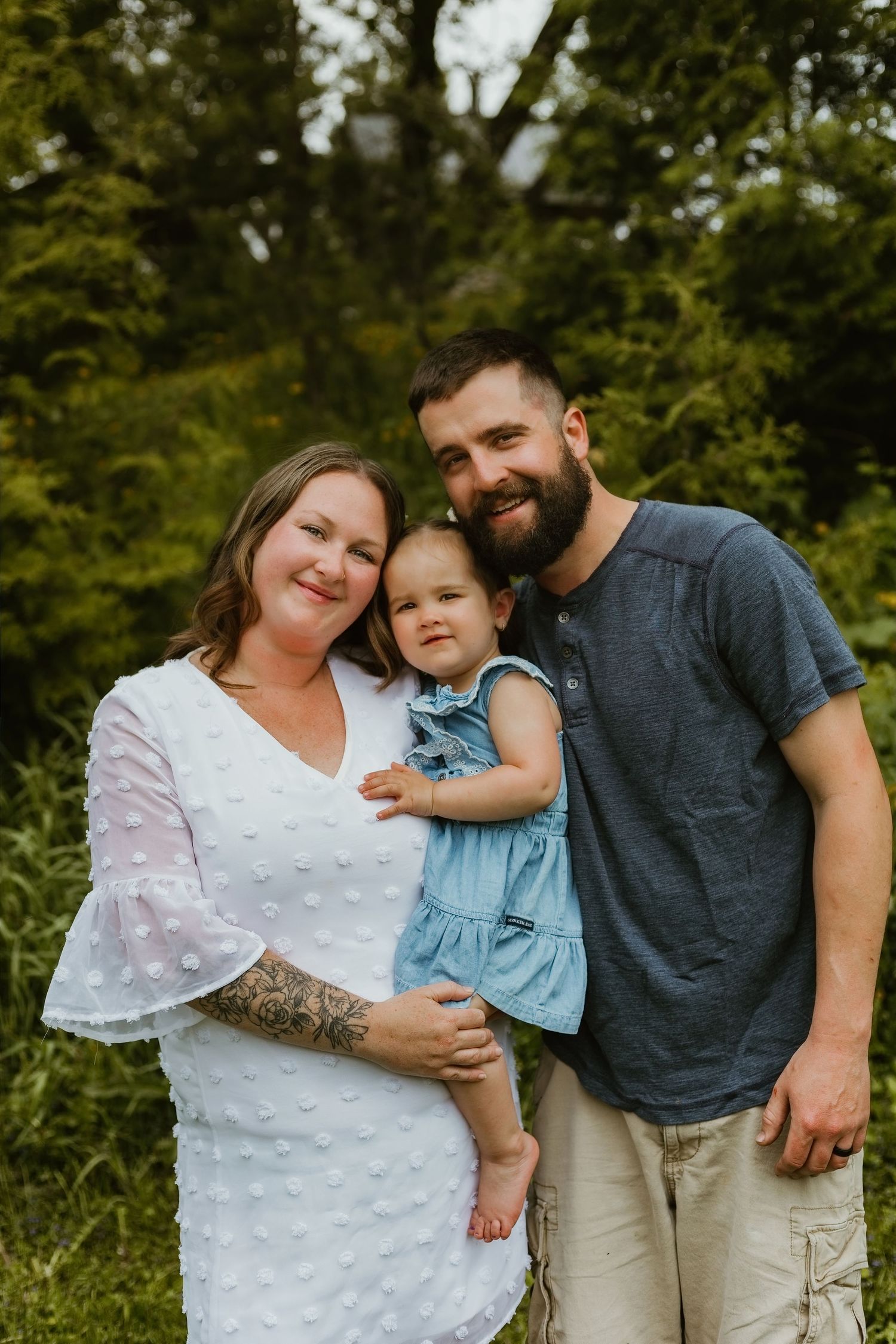 A smiling family of three poses outdoors, with a woman in white, bearded man in navy, and baby girl in denim dress.