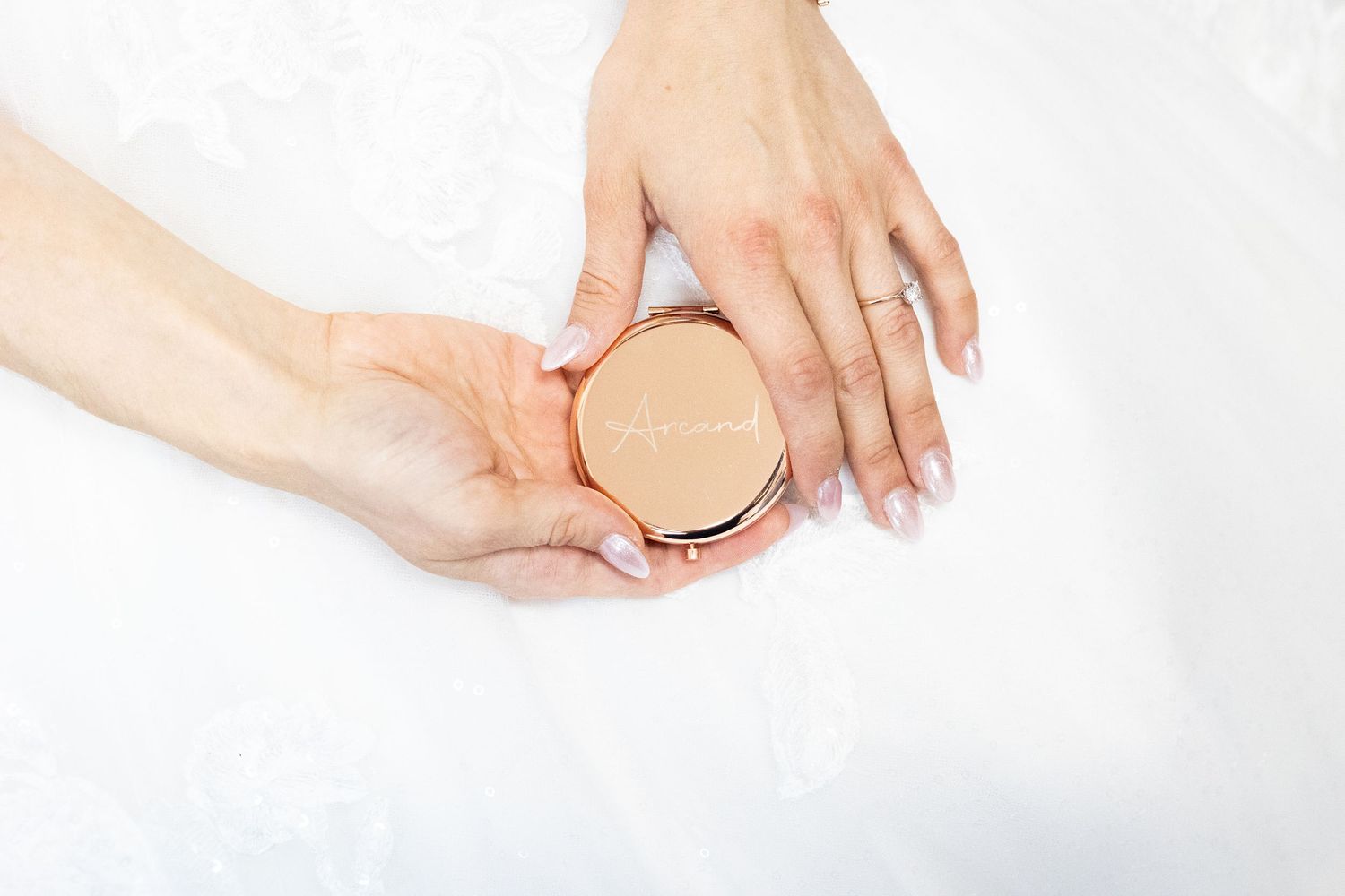 Close-up of bride's hands holding a rose gold compact mirror against white wedding dress fabric.