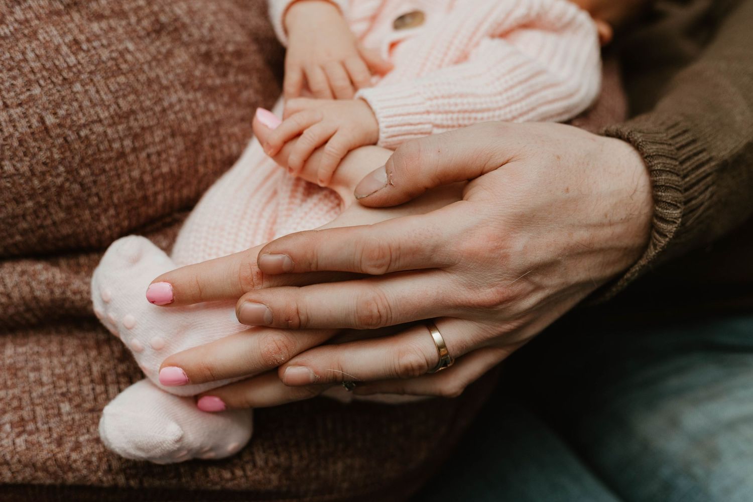 Close-up of mother's manicured hands holding newborn baby's pink-socked feet on a couch.