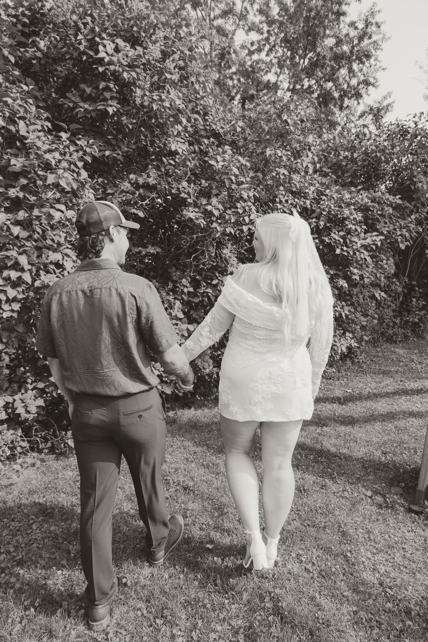 Black and white photo of bride and groom holding hands walking through lush garden greenery.