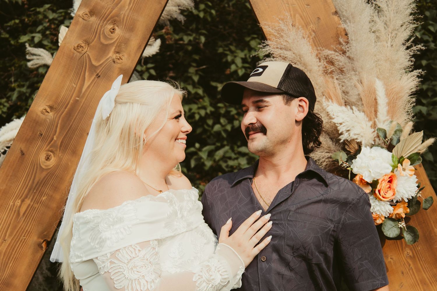 Smiling bride and groom pose before wooden triangle arch with pampas grass decor.