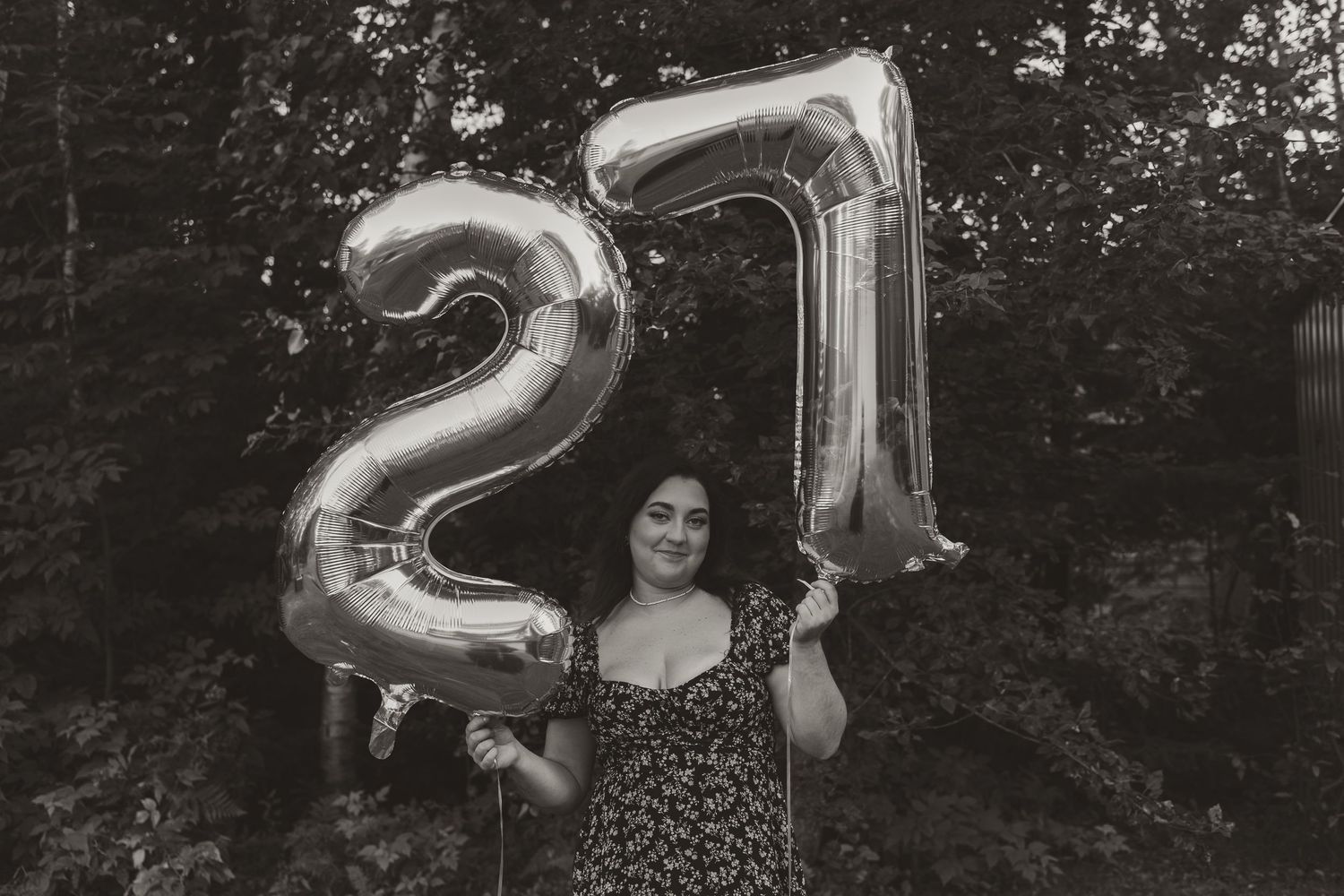 Woman holding large silver number 27 balloons outdoors, celebrating her 27th birthday in a floral dress.