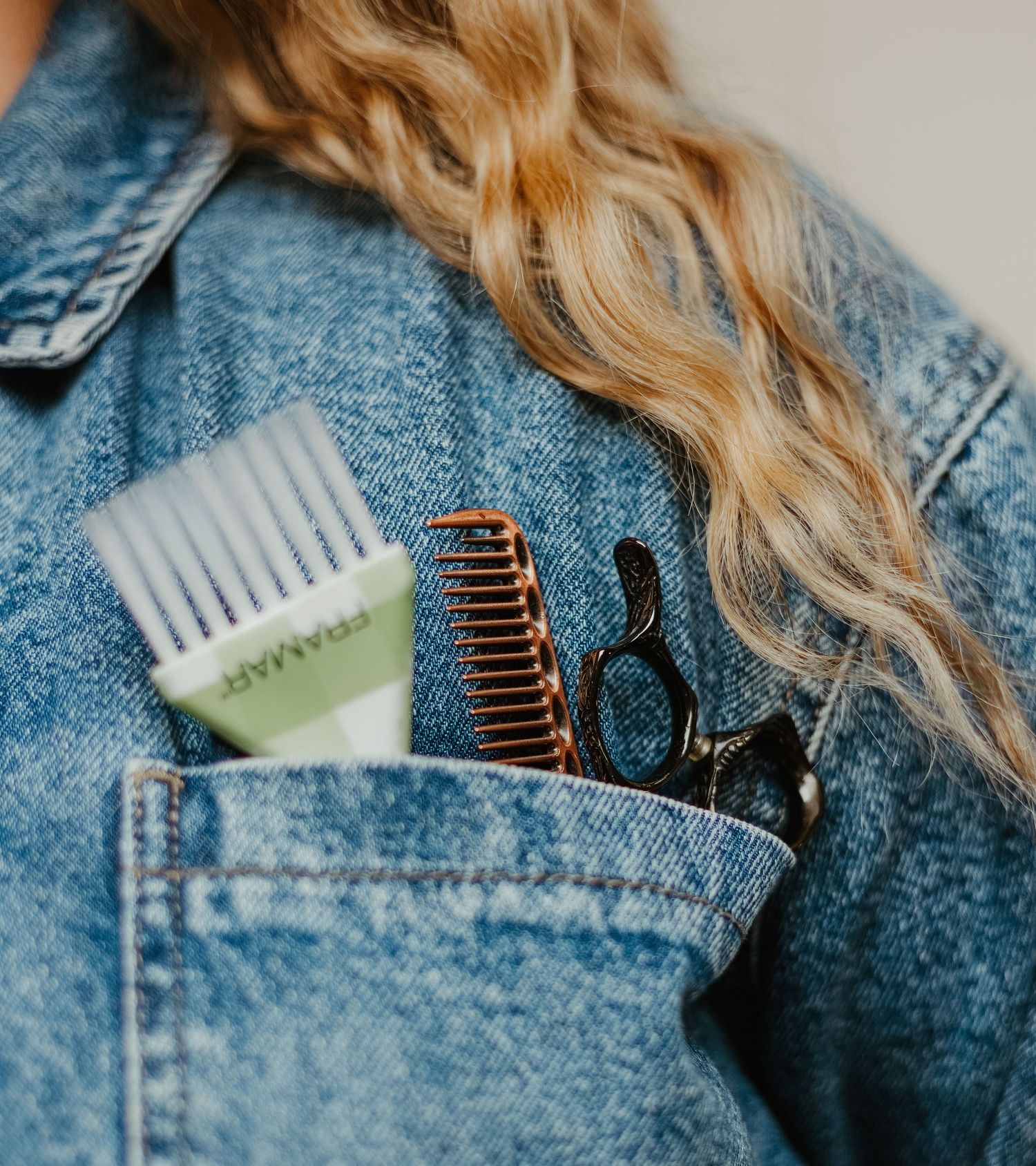 Blonde woman in denim jacket with hair tools in pocket, including comb, brush, and scissors.