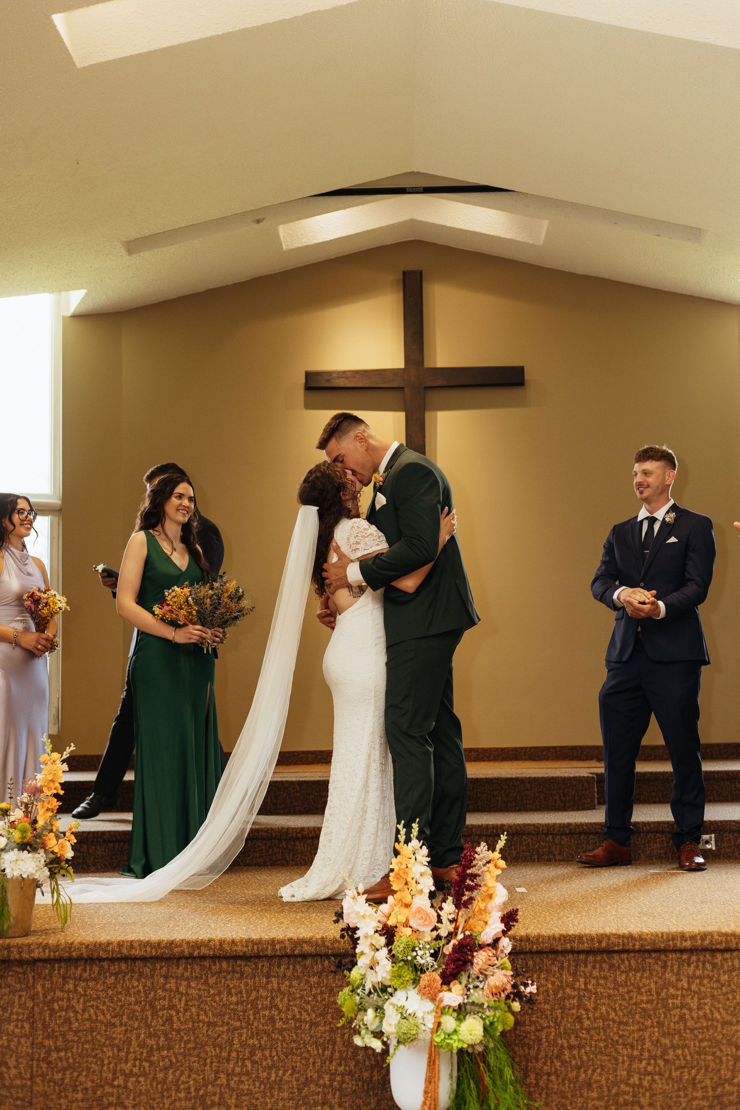 Bride and groom share first kiss at church altar with cross backdrop, flanked by wedding party in green and navy attire.