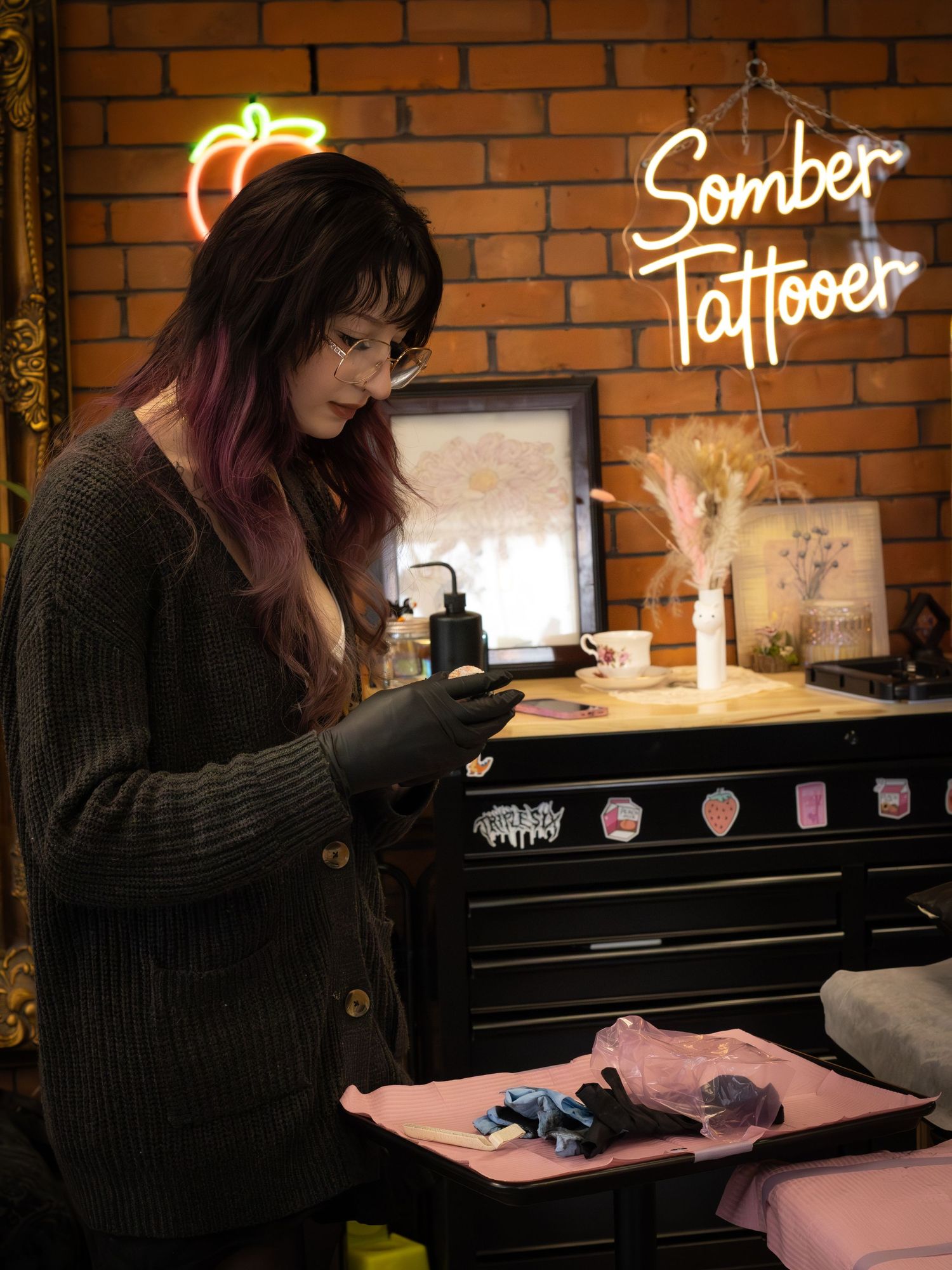 Tattooed woman in cardigan preparing tattoo equipment at Somber Tattooer studio.