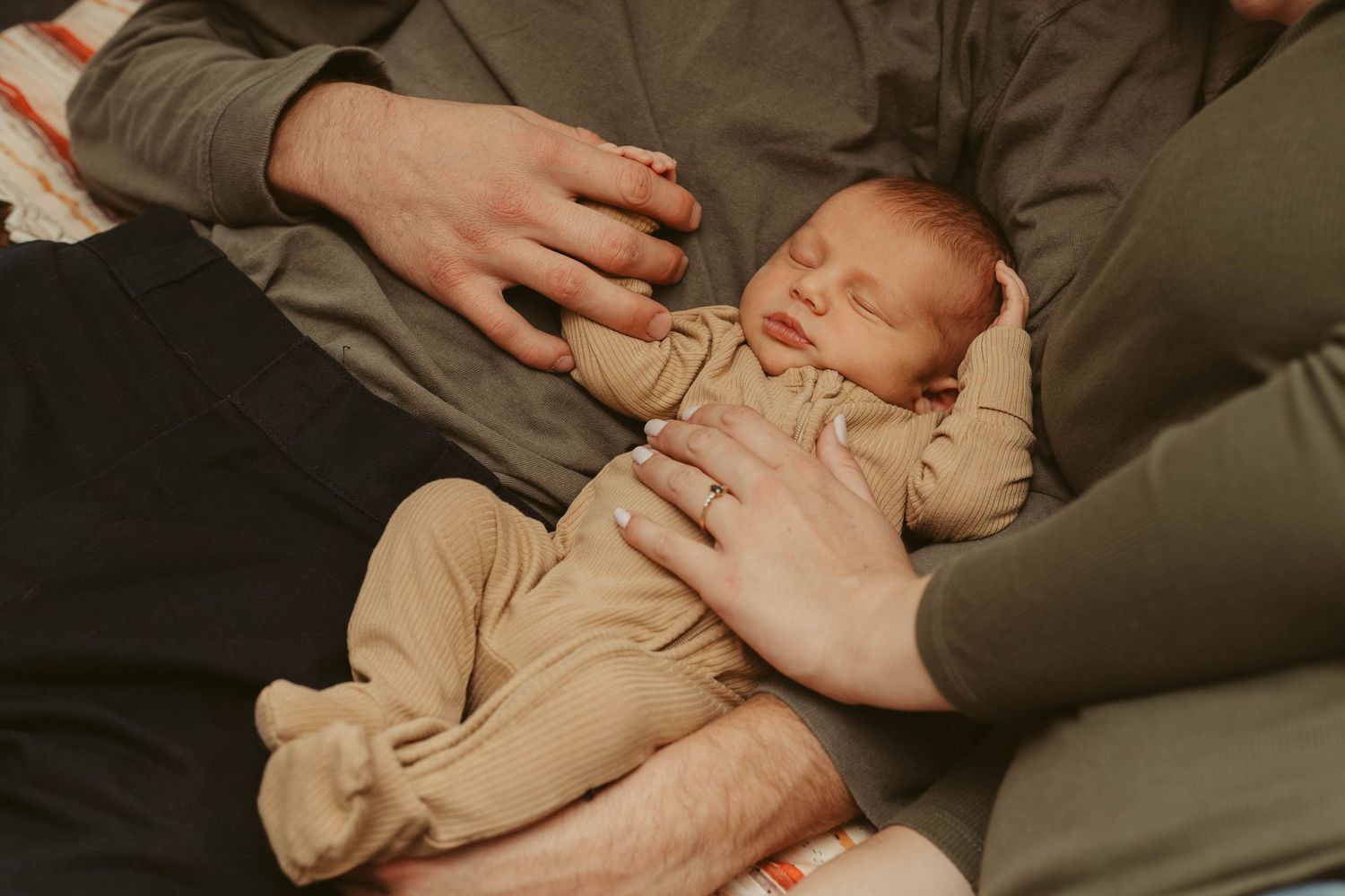 Sleeping newborn baby cradled in parents' laps with gentle hands resting on chest.