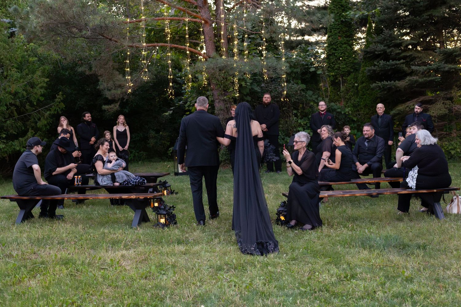 Gothic outdoor wedding ceremony with guests seated on wooden benches surrounded by trees and string lights.