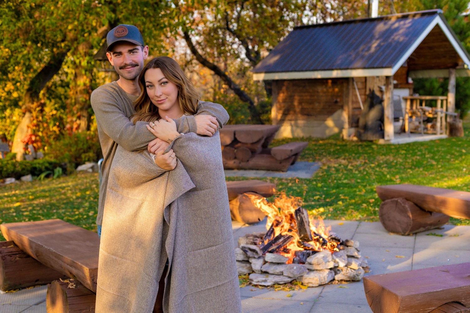 Couple wrapped in blanket embracing warmly beside outdoor campfire near rustic cabin.