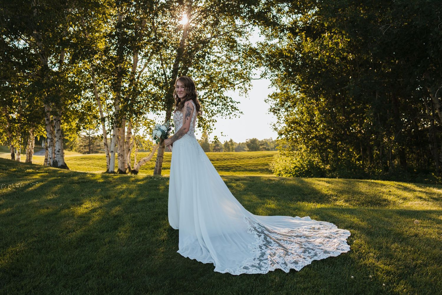 Bride in white gown with long train stands in sunlit garden among trees, holding bouquet on wedding day.