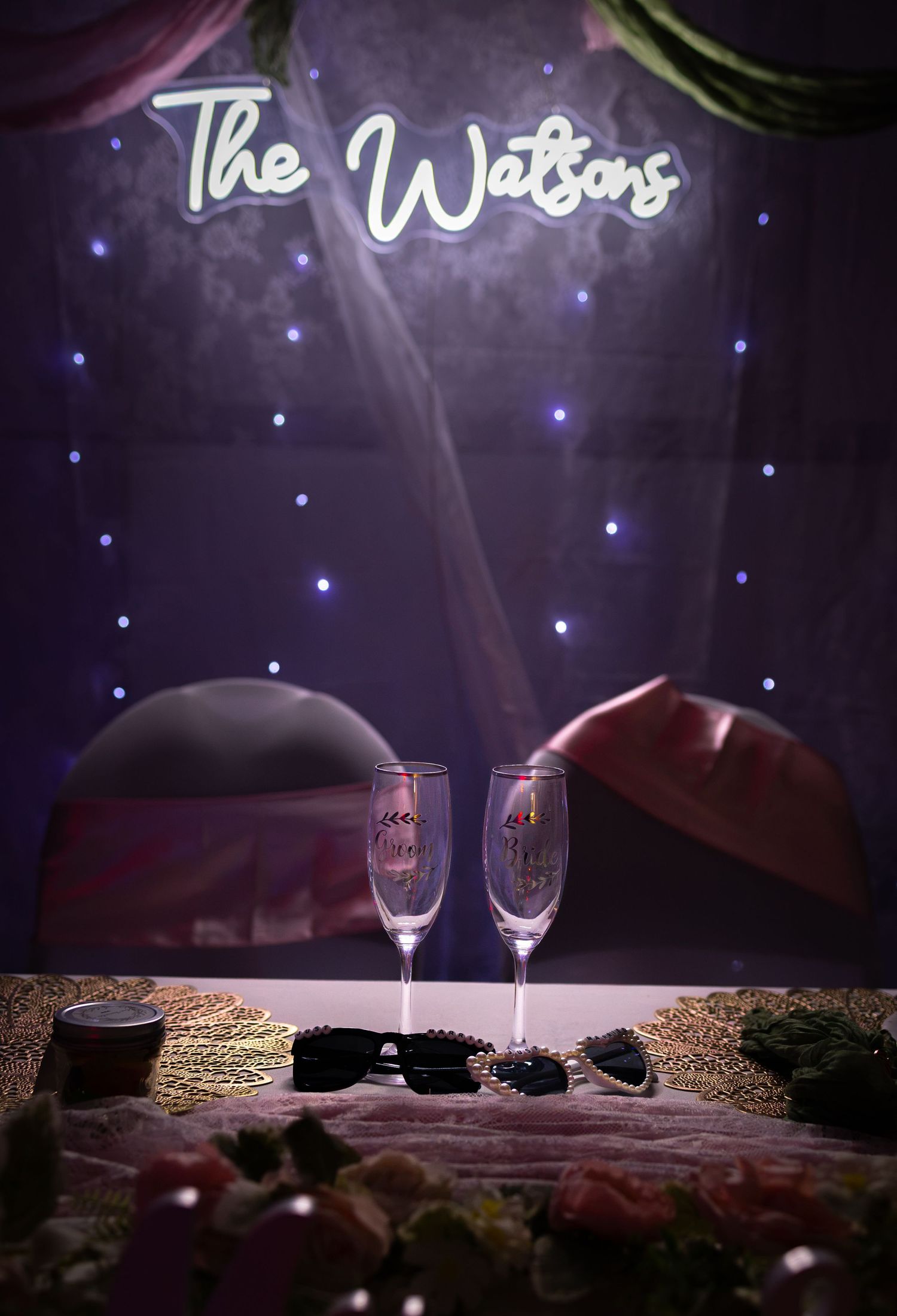 Two champagne flutes on a decorated table under a glowing neon sign reading The Watsons at a wedding reception.