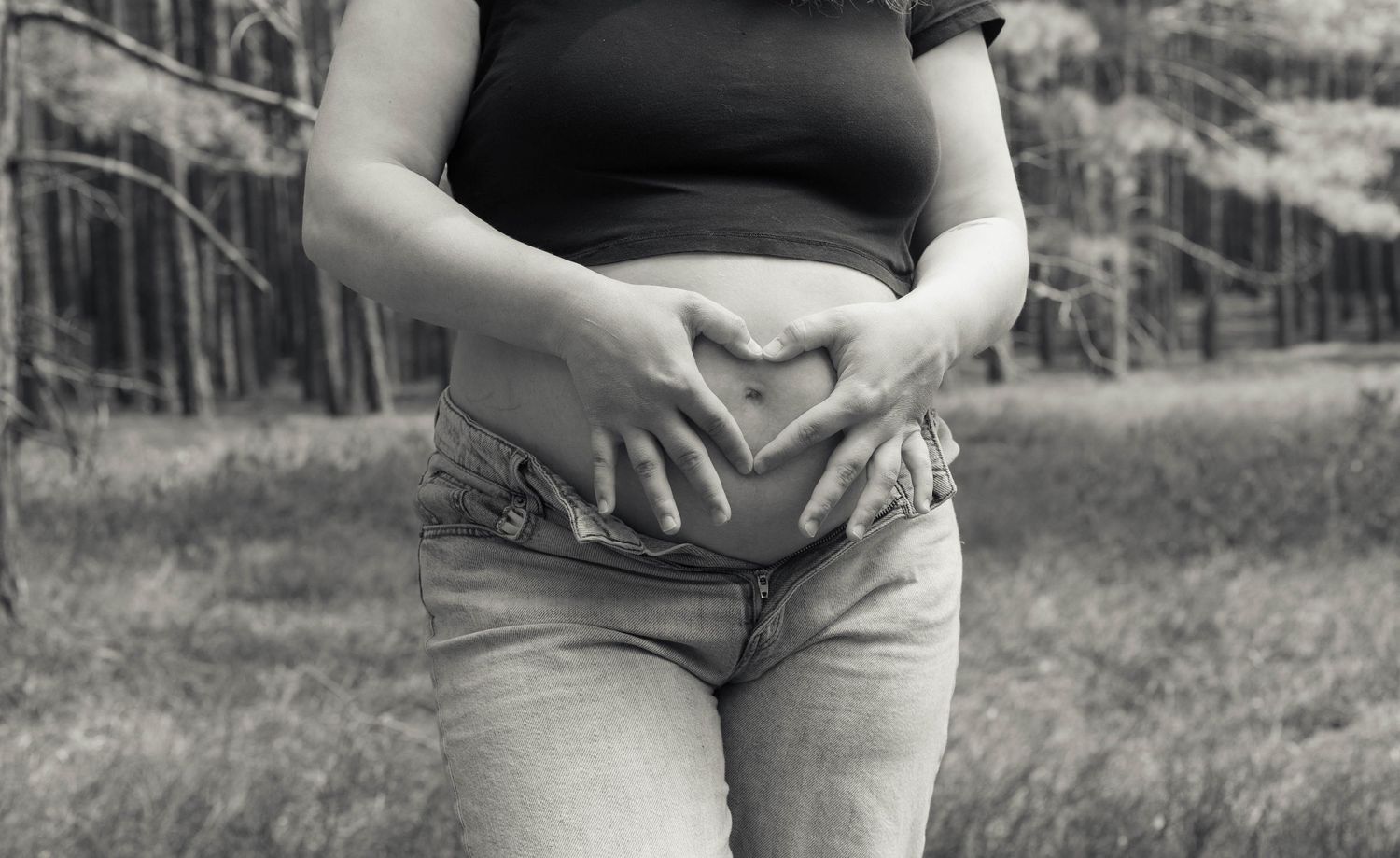 Black and white photo of pregnant woman making heart shape with hands on baby bump outdoors.