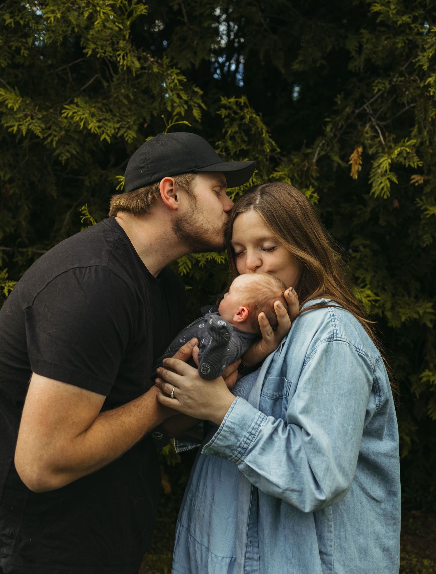 A couple shares a tender kiss while cradling their newborn baby outdoors near lush green trees.