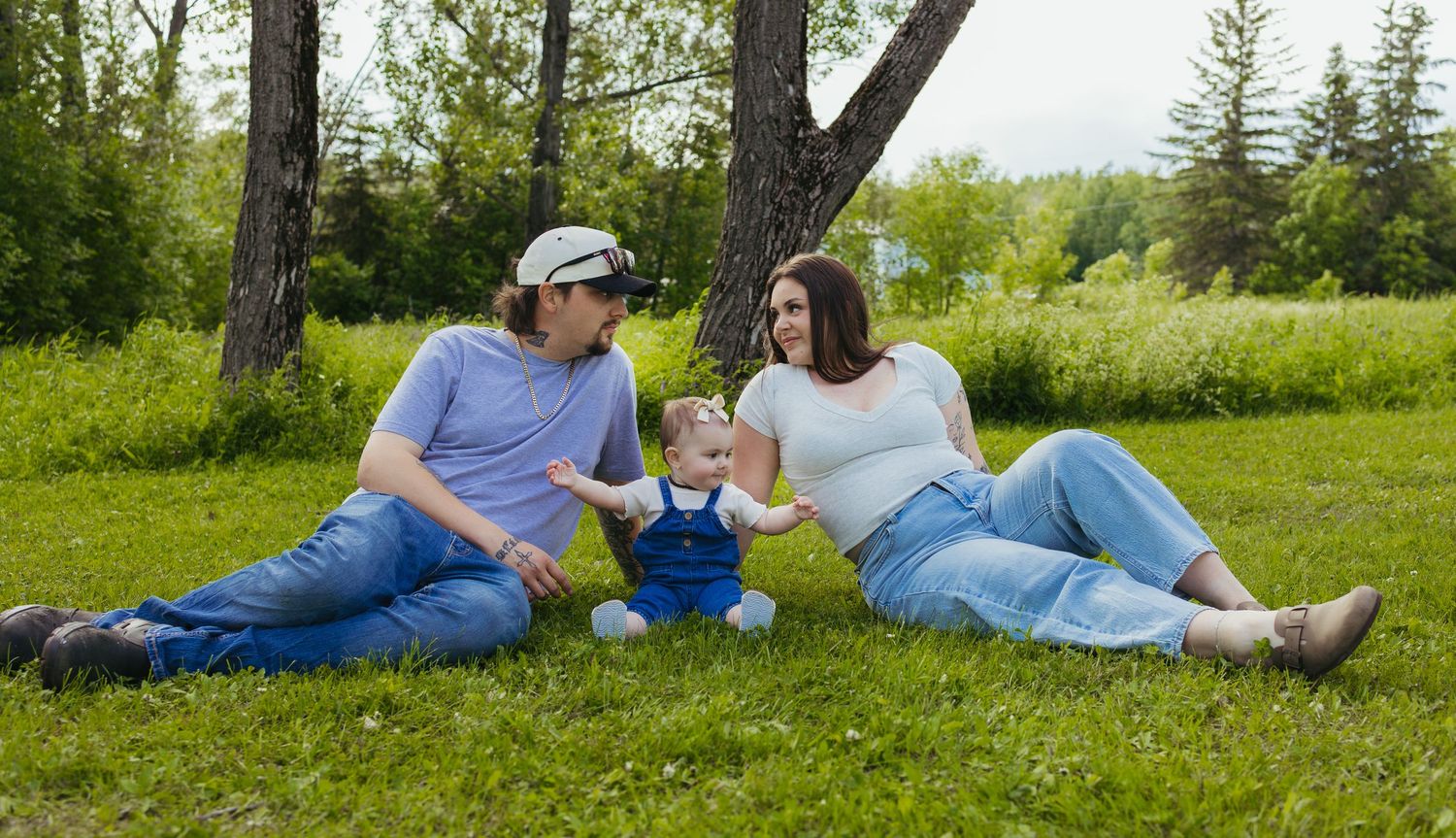 Family sitting on grass under trees, parents smiling at baby in blue outfit during outdoor maternity photo session.
