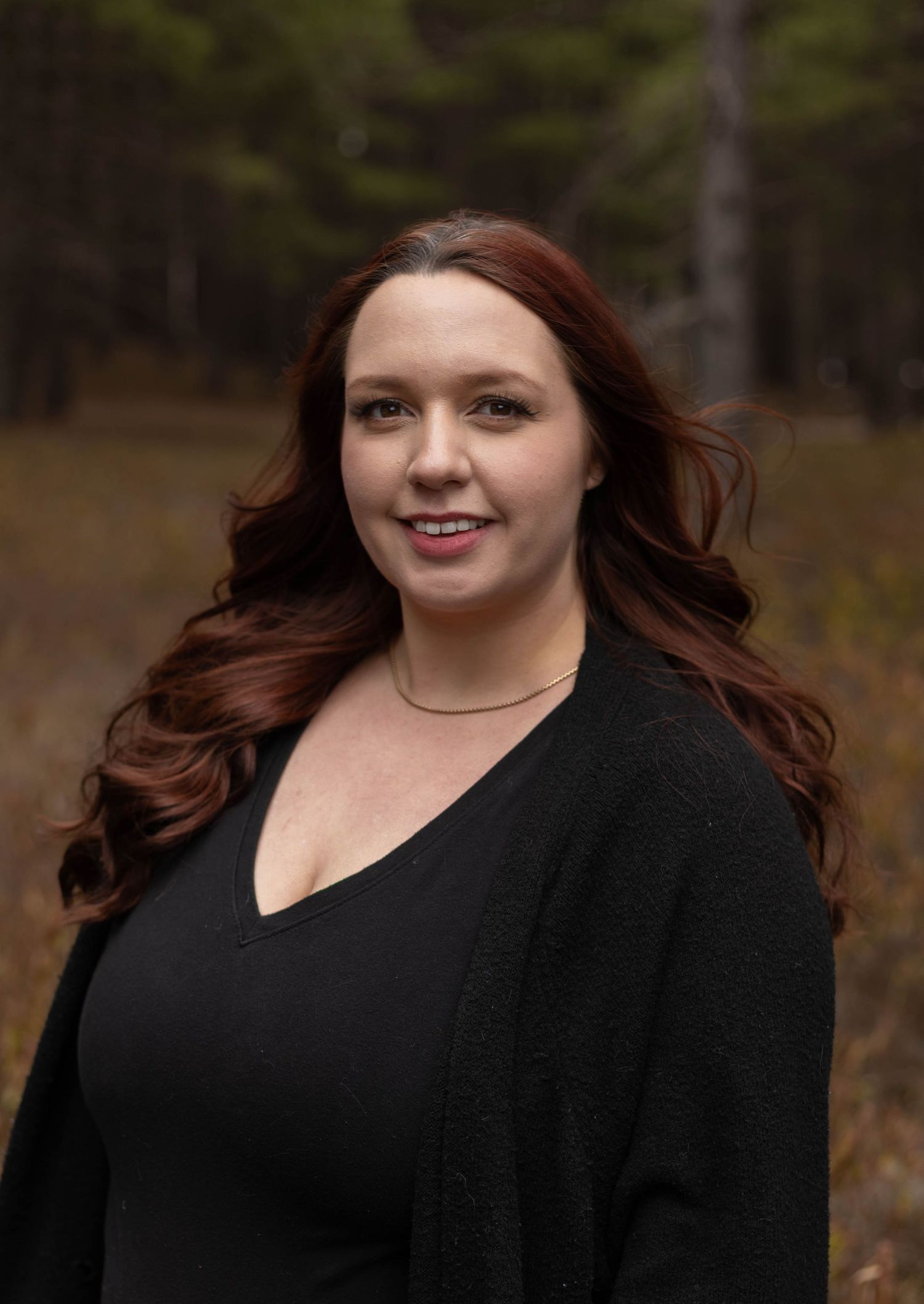 Woman with red hair wearing black cardigan smiling outdoors in forest setting, portrait photo.