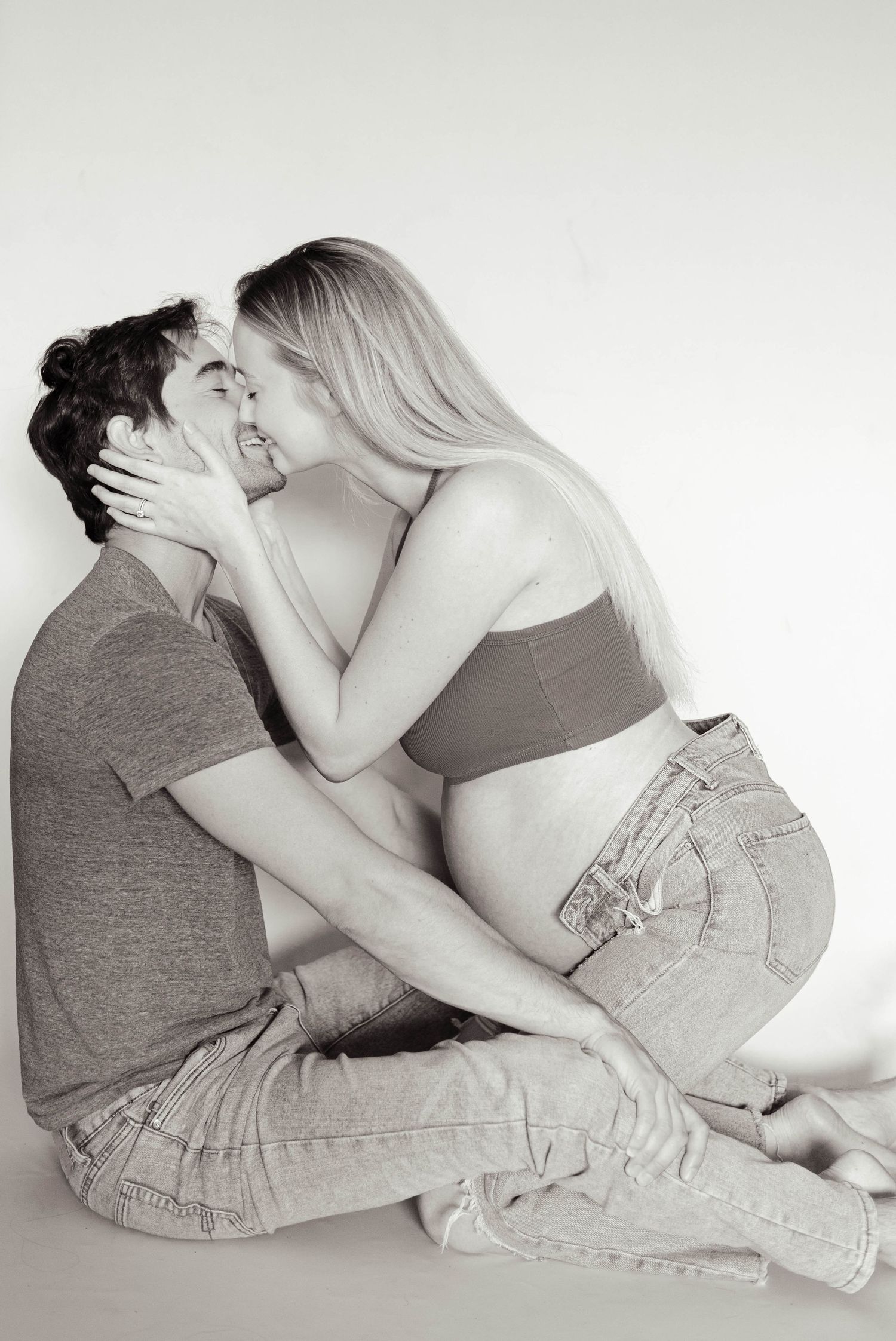 Black and white photo of pregnant couple sharing a kiss while sitting on floor together.