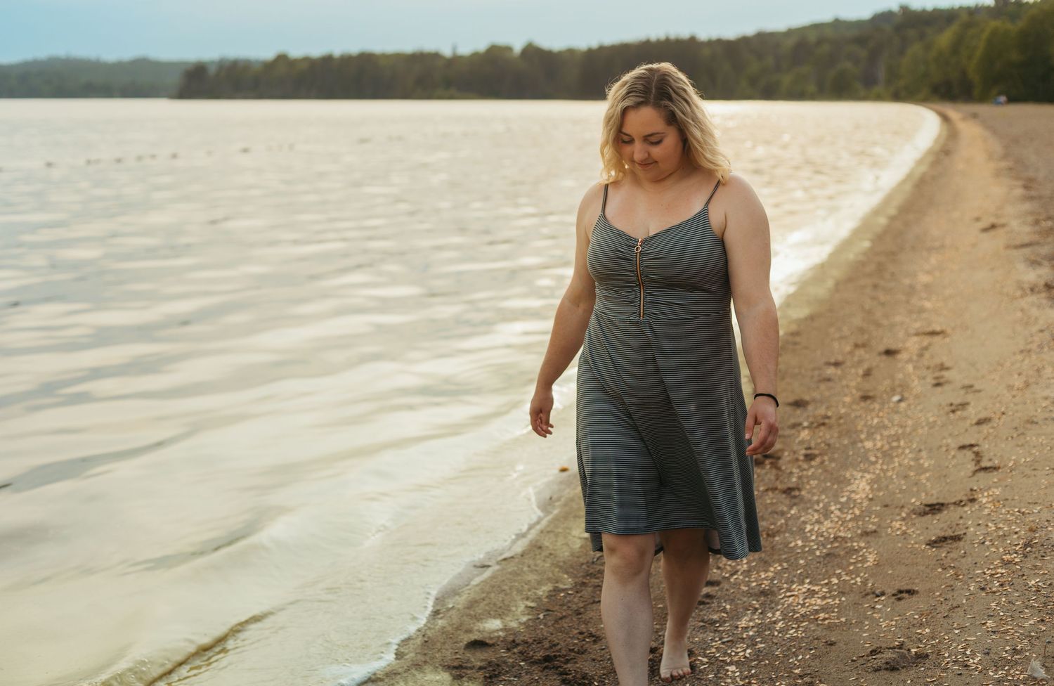 Woman in striped sundress walking along sandy beach shoreline near calm water.