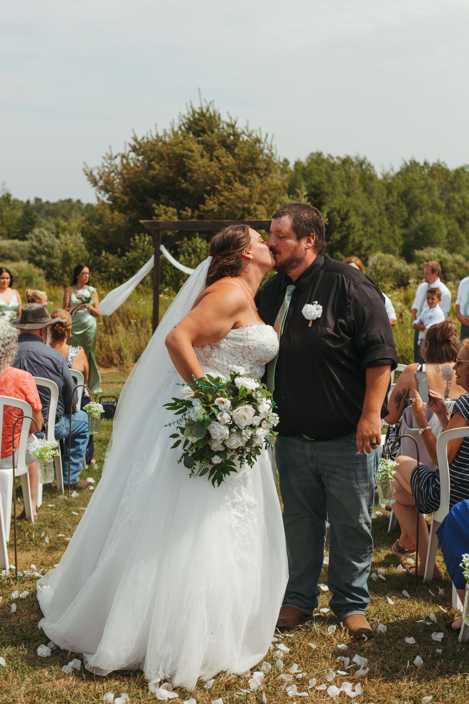 Bride and groom sharing first kiss at outdoor wedding ceremony with guests seated on lawn.