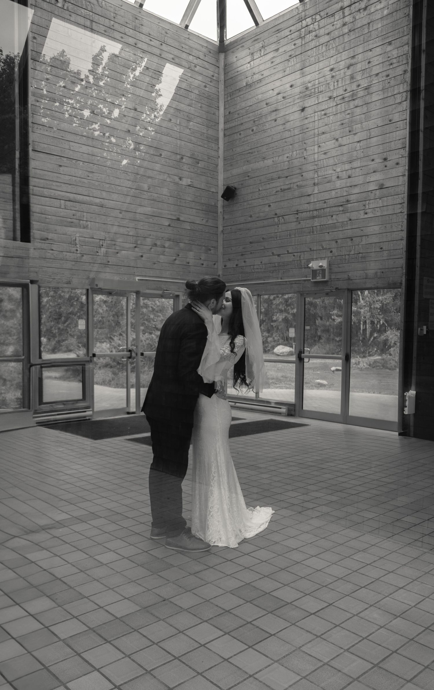 Black and white photo of bride and groom sharing a kiss in a modern building lobby with tile floors.