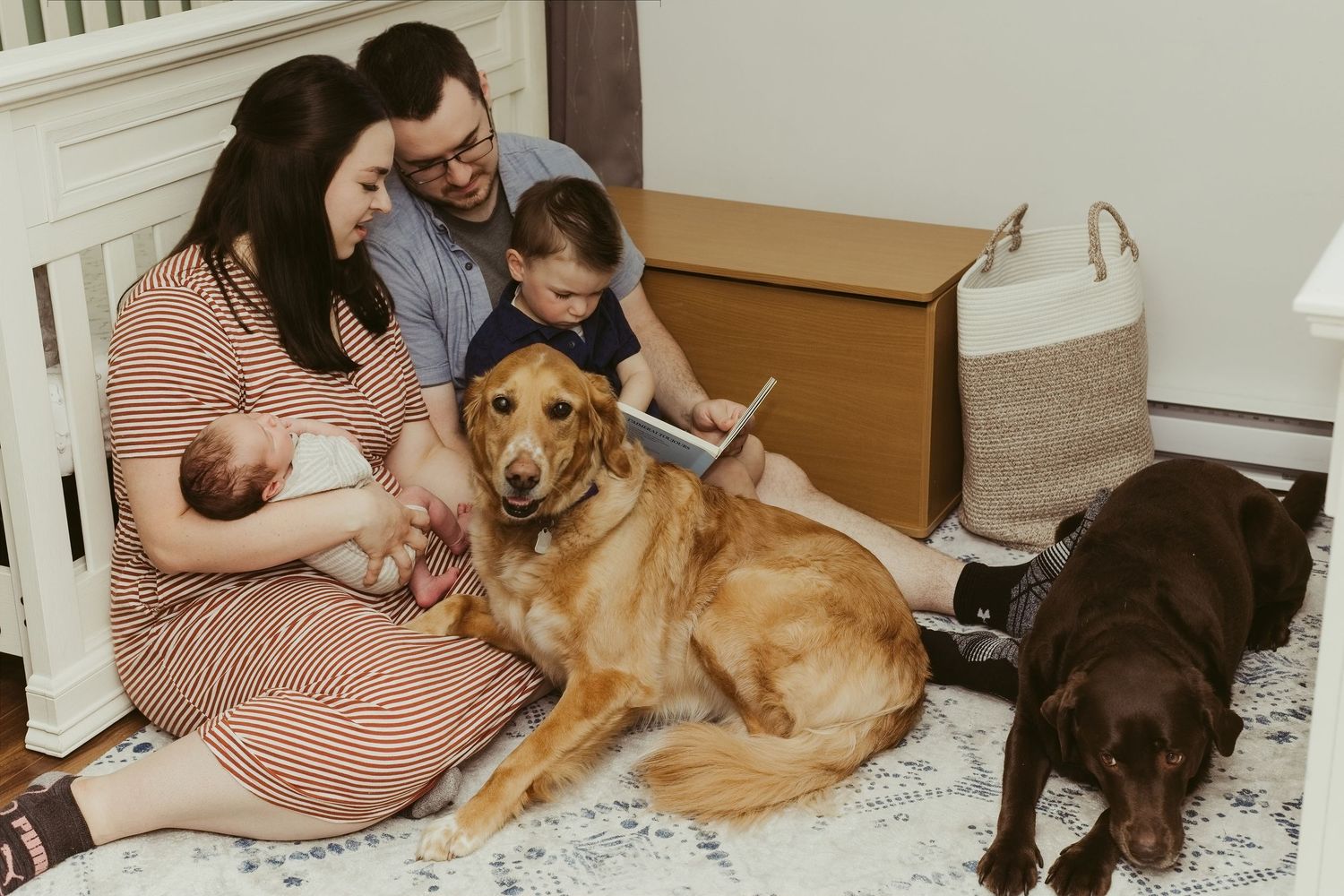Family with newborn, toddler, and two dogs sitting on floor by fireplace in cozy living room.