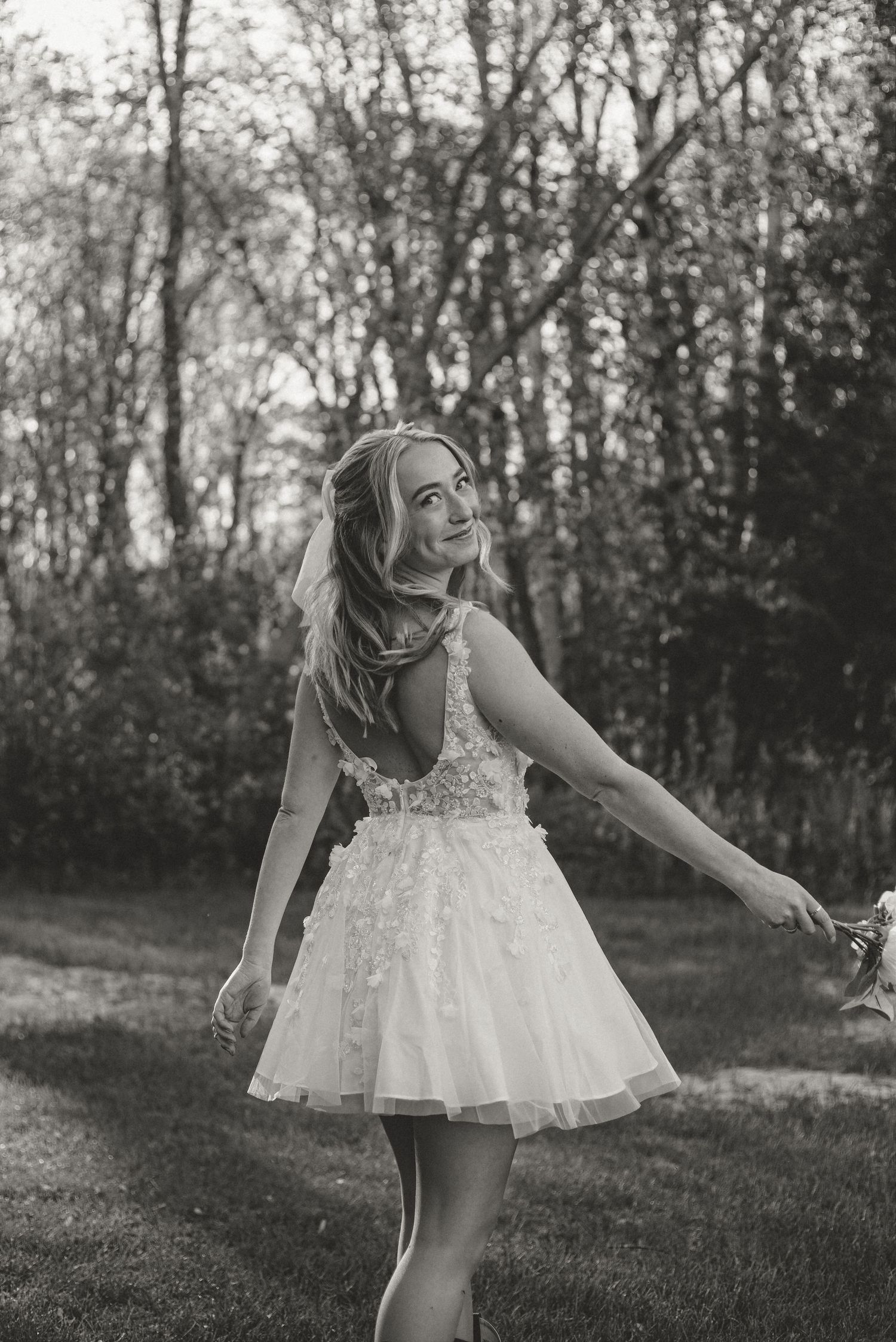 Black and white photo of a smiling bride in a short floral wedding dress twirling outdoors with trees in the background.