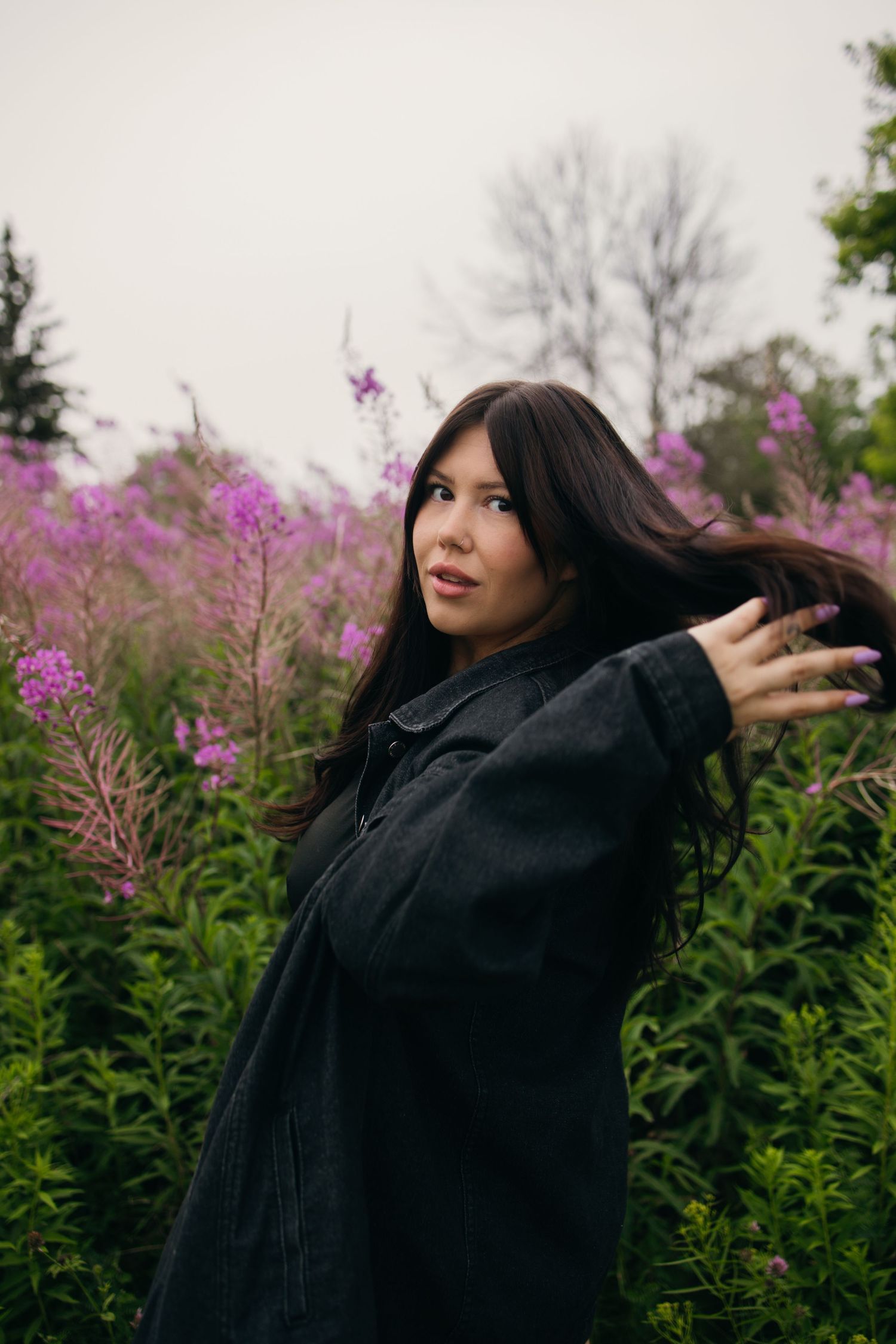 Young woman with long dark hair in black coat posing among vibrant pink wildflowers in a lush green field.