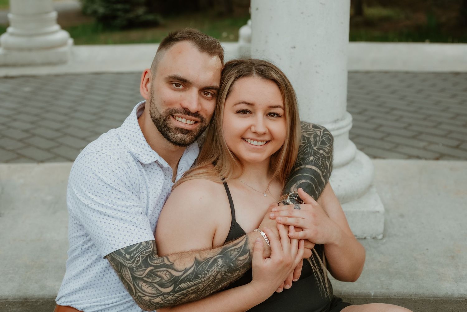 Happy couple posing together outdoors, man with tattoos embracing smiling woman near white column.
