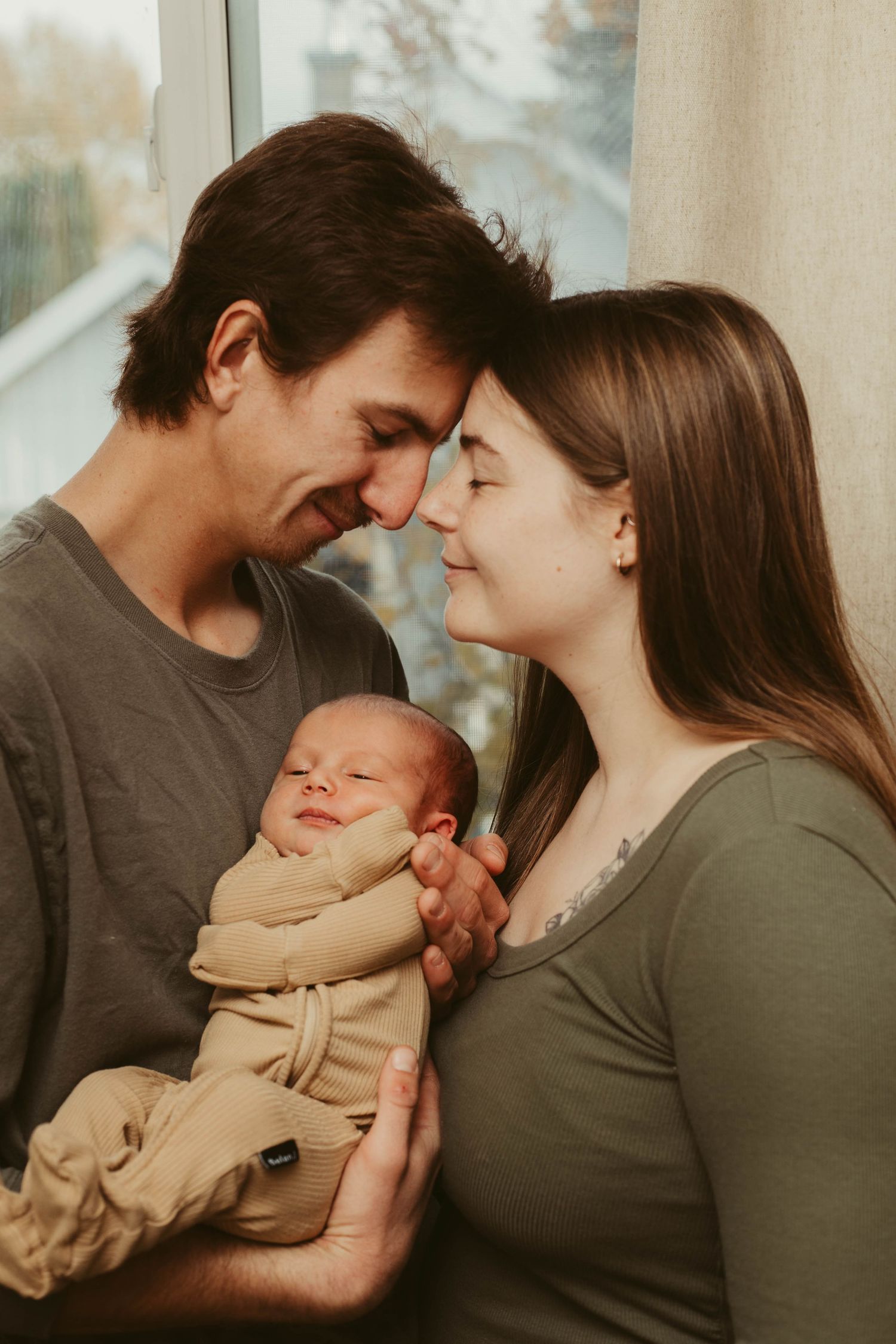 New parents touching foreheads tenderly while holding their sleeping newborn baby by a window.