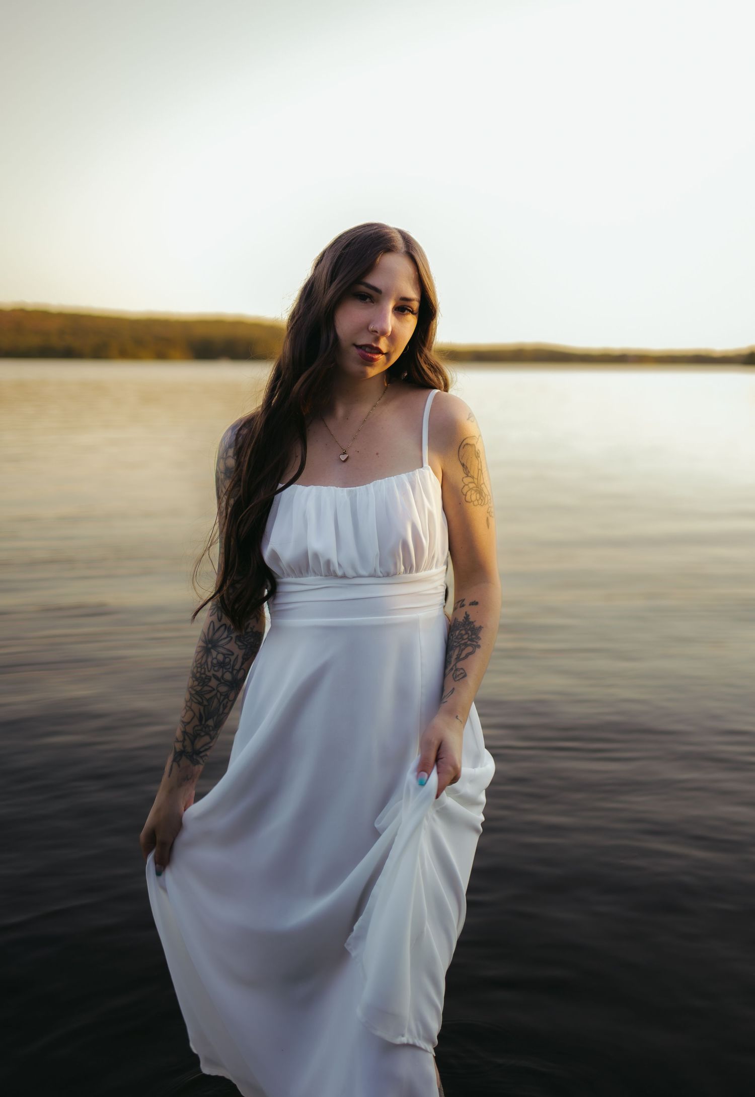 A woman with long dark hair and tattoos poses in a white sundress standing in a calm lake at golden hour.