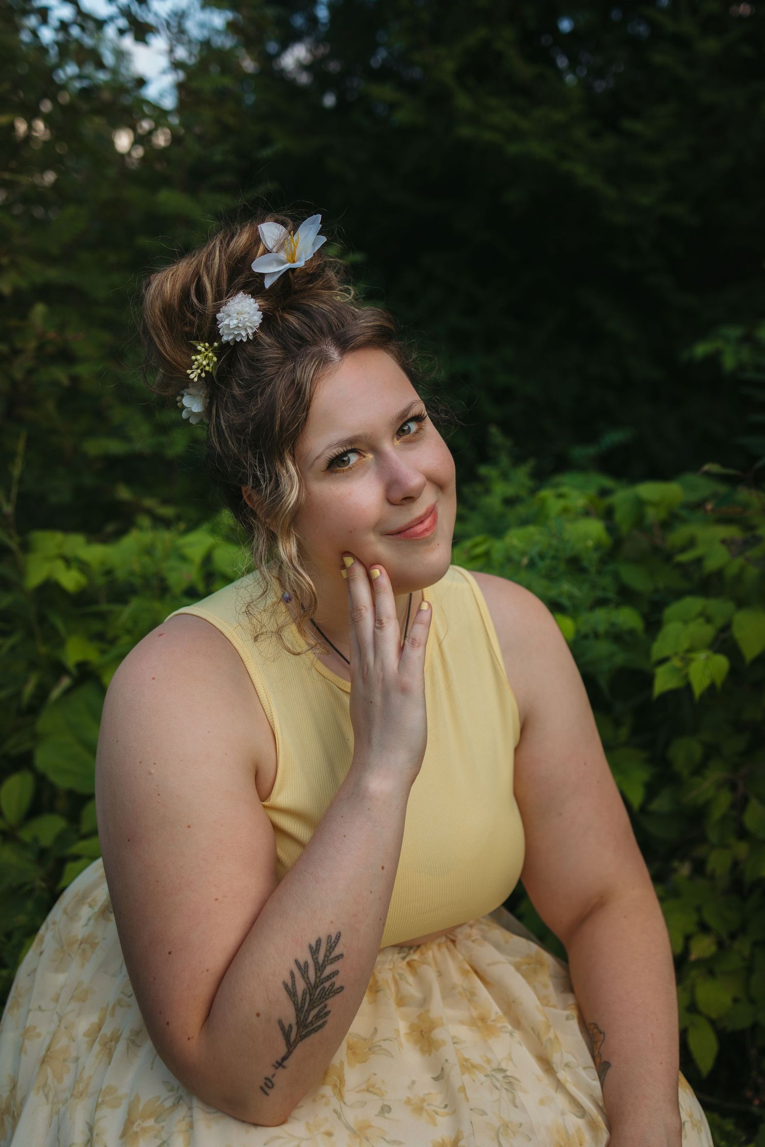 Woman in yellow dress with floral hair accessories poses outdoors among lush green foliage, showing arm tattoo.