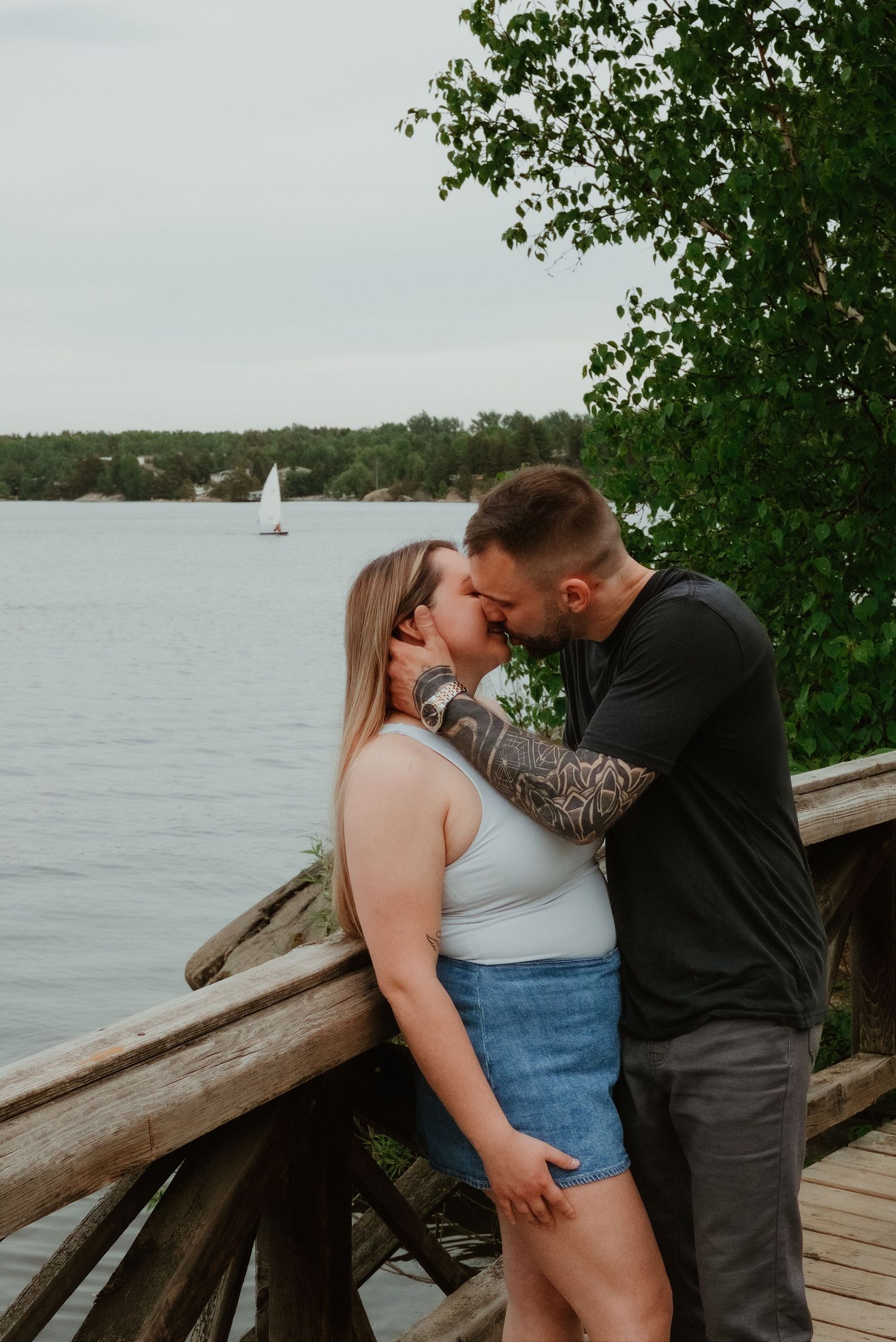 Couple sharing a romantic kiss on a wooden dock by a lake, man with tattoo sleeve embracing woman in white top.