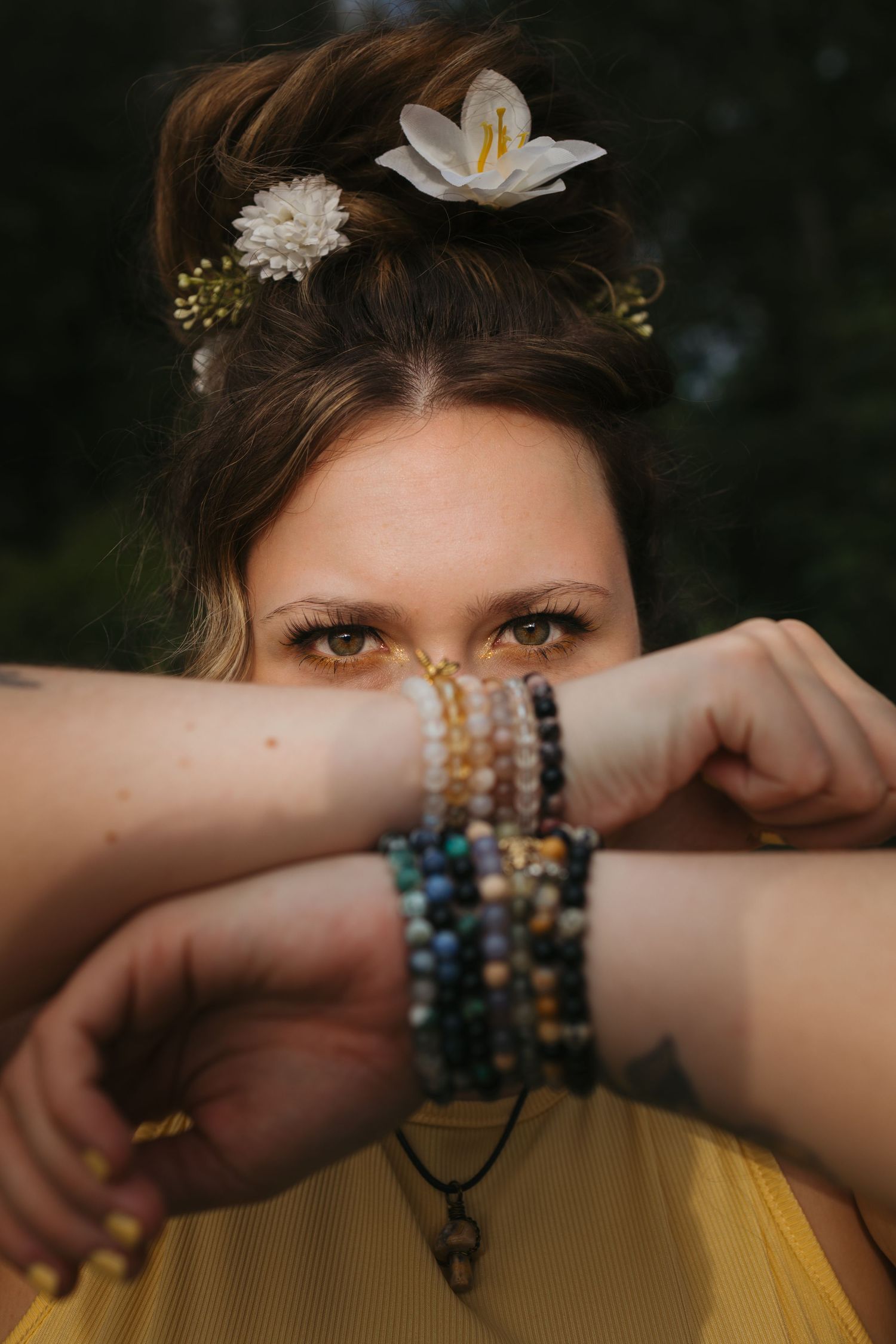 Woman with flowers in her hair wearing colorful beaded bracelets, covering her face with her wrists.