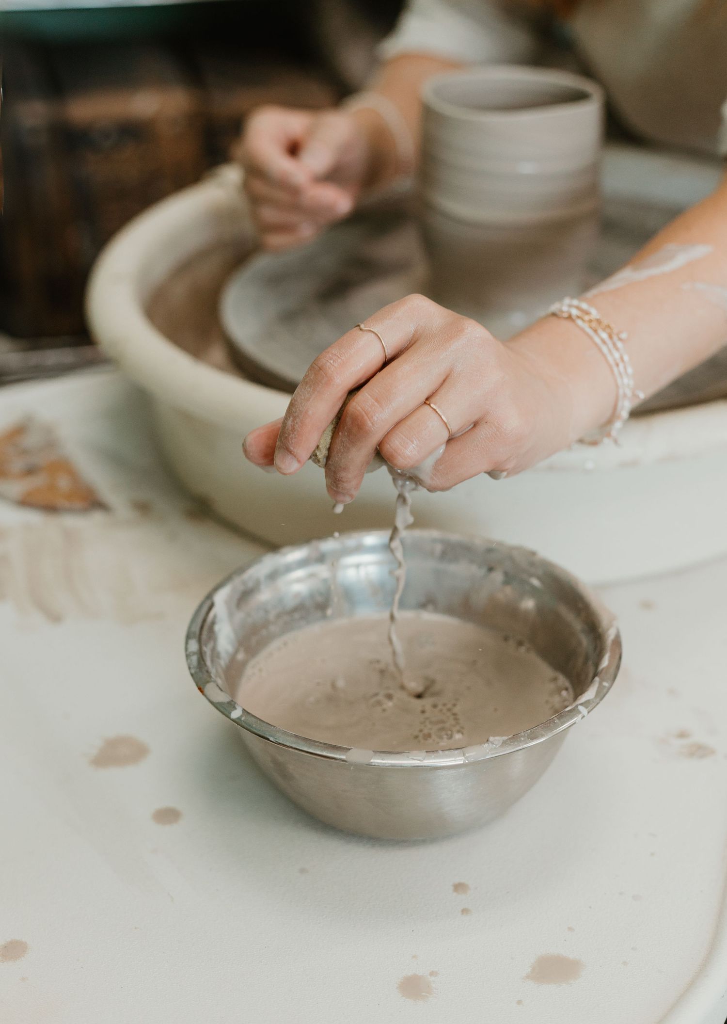 Hands dripping water into a metal bowl of clay slip while working on a pottery wheel in a ceramic studio.