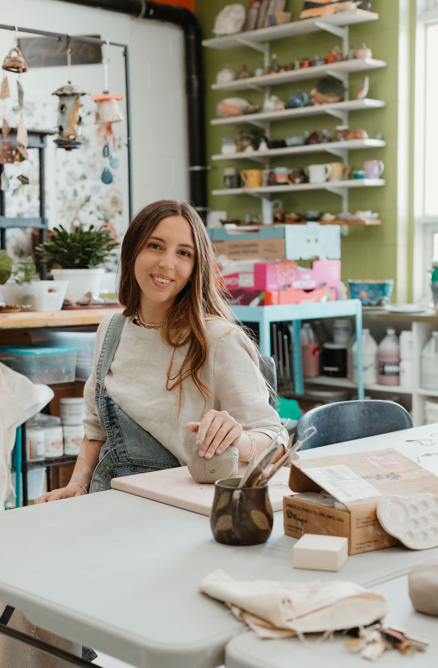 A smiling woman in overalls sits at a worktable in a colorful pottery studio surrounded by shelves of ceramics.