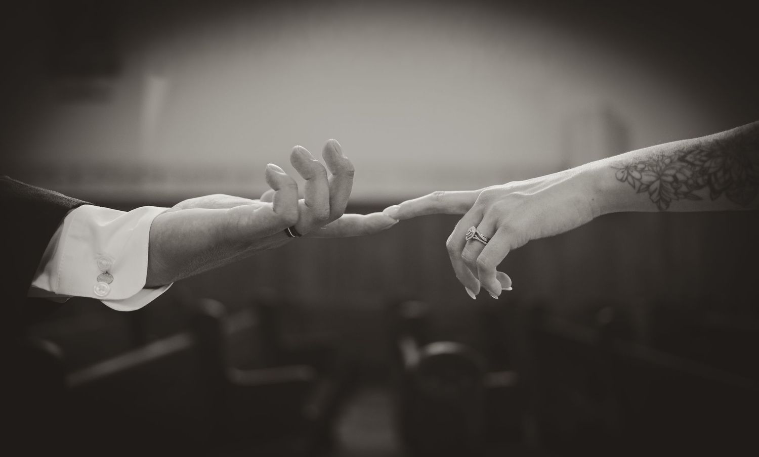 Black and white photo of two hands almost touching, one with a wedding ring and tattoo, evoking romance.