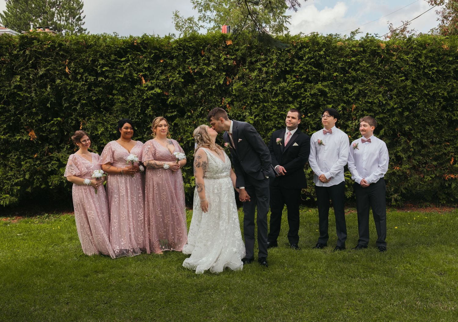 Wedding party poses outdoors with bride and groom kissing, bridesmaids in pink gowns and groomsmen in suits before a green hedge.