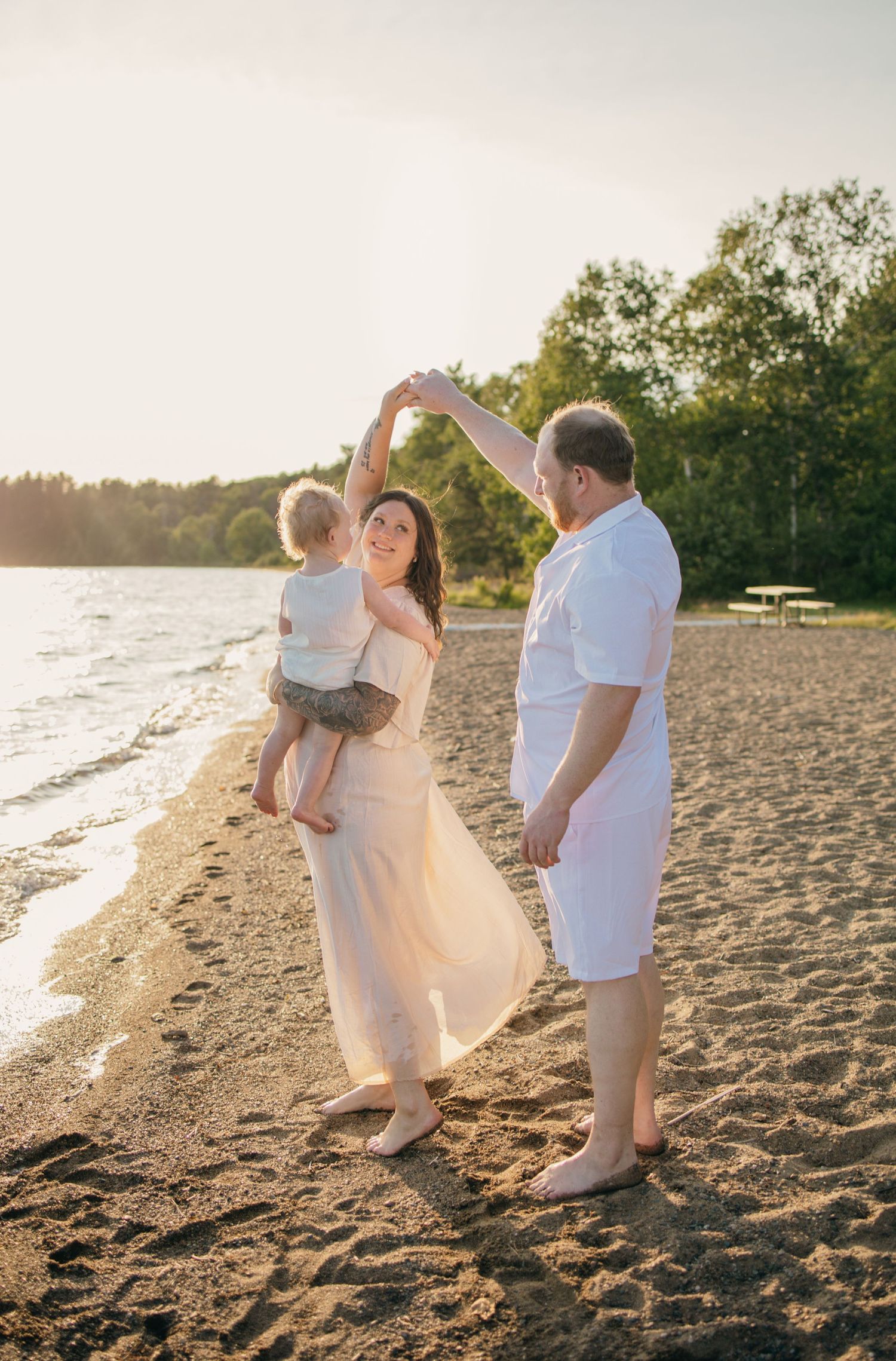 Family dancing on sandy beach at golden sunset, mother in white dress holding toddler while father twirls her hand.