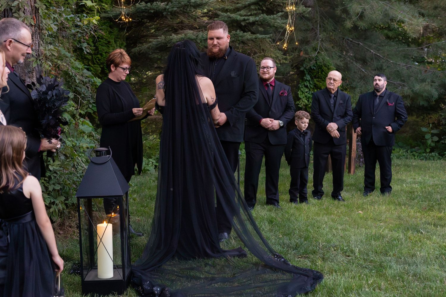 Bride in black gown with long veil standing at outdoor gothic wedding ceremony with groom and groomsmen.