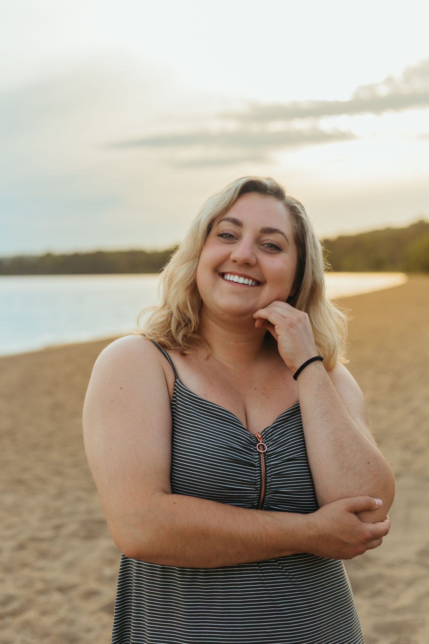 Smiling blonde woman in striped sundress posing on sandy beach at golden hour sunset.