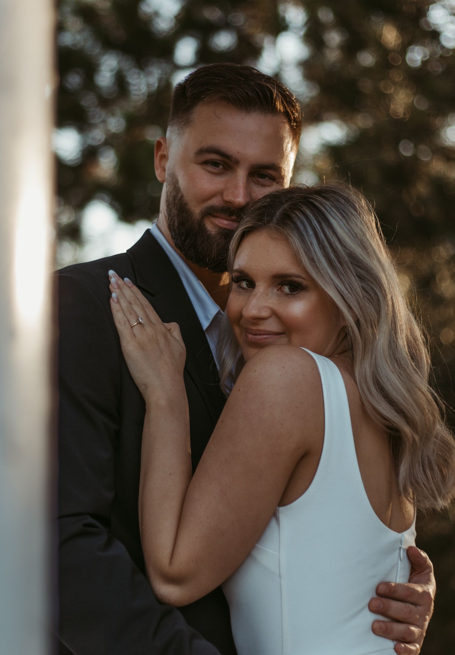 Engaged couple posing together, woman in white dress showing ring, man in dark suit, outdoor portrait.